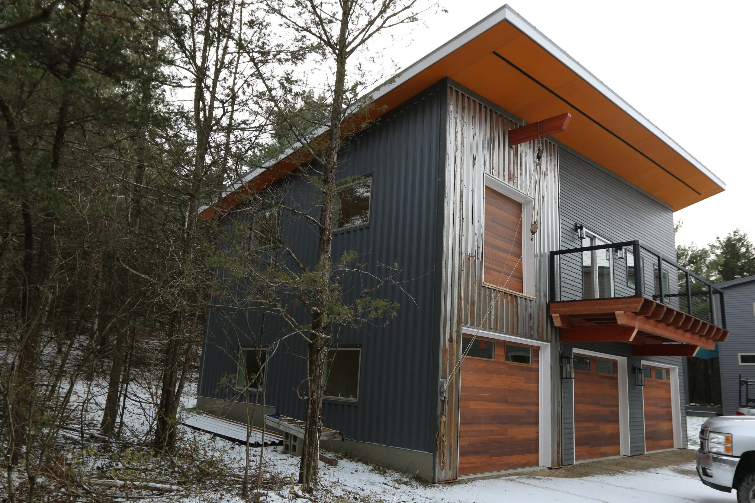 Modern two-story house with mixed metal and wood exterior, garage doors, small front balcony, surrounded by trees, with a driveway and a truck parked outside.