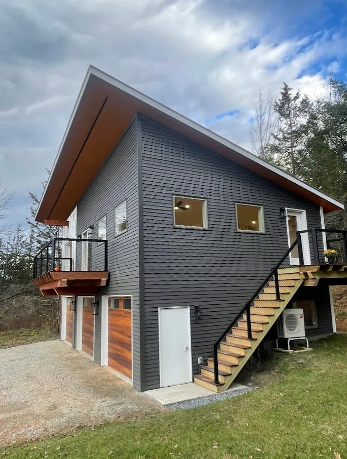 Modern two-story house with dark gray siding, wooden garage doors, and an exterior staircase leading to a second-floor entrance, set against a clouded sky and surrounded by trees.