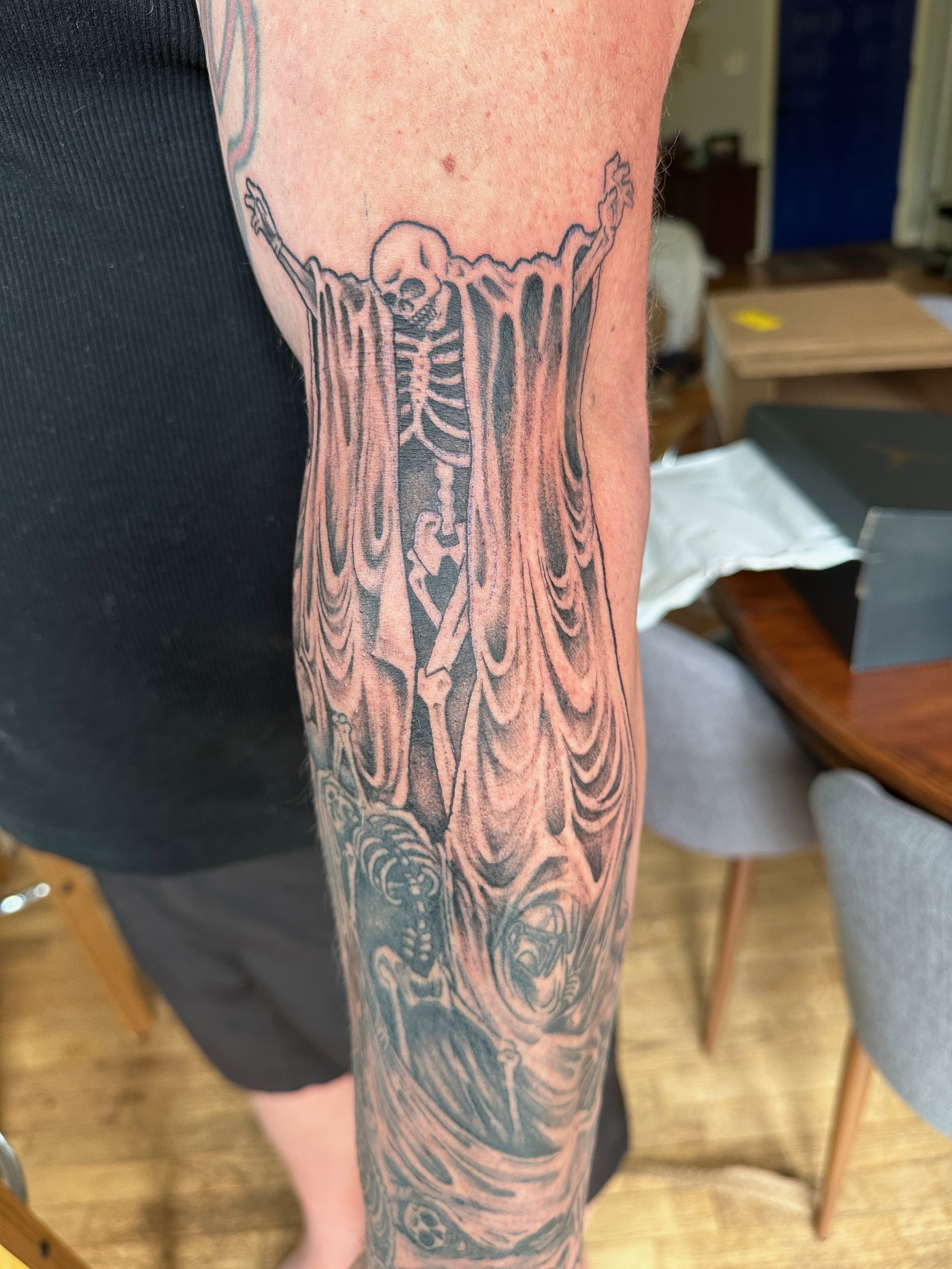 Tattoo of a skeleton with outstretched arms and a flowing cloak on a person's arm. Tattoo designed by Robin Reece