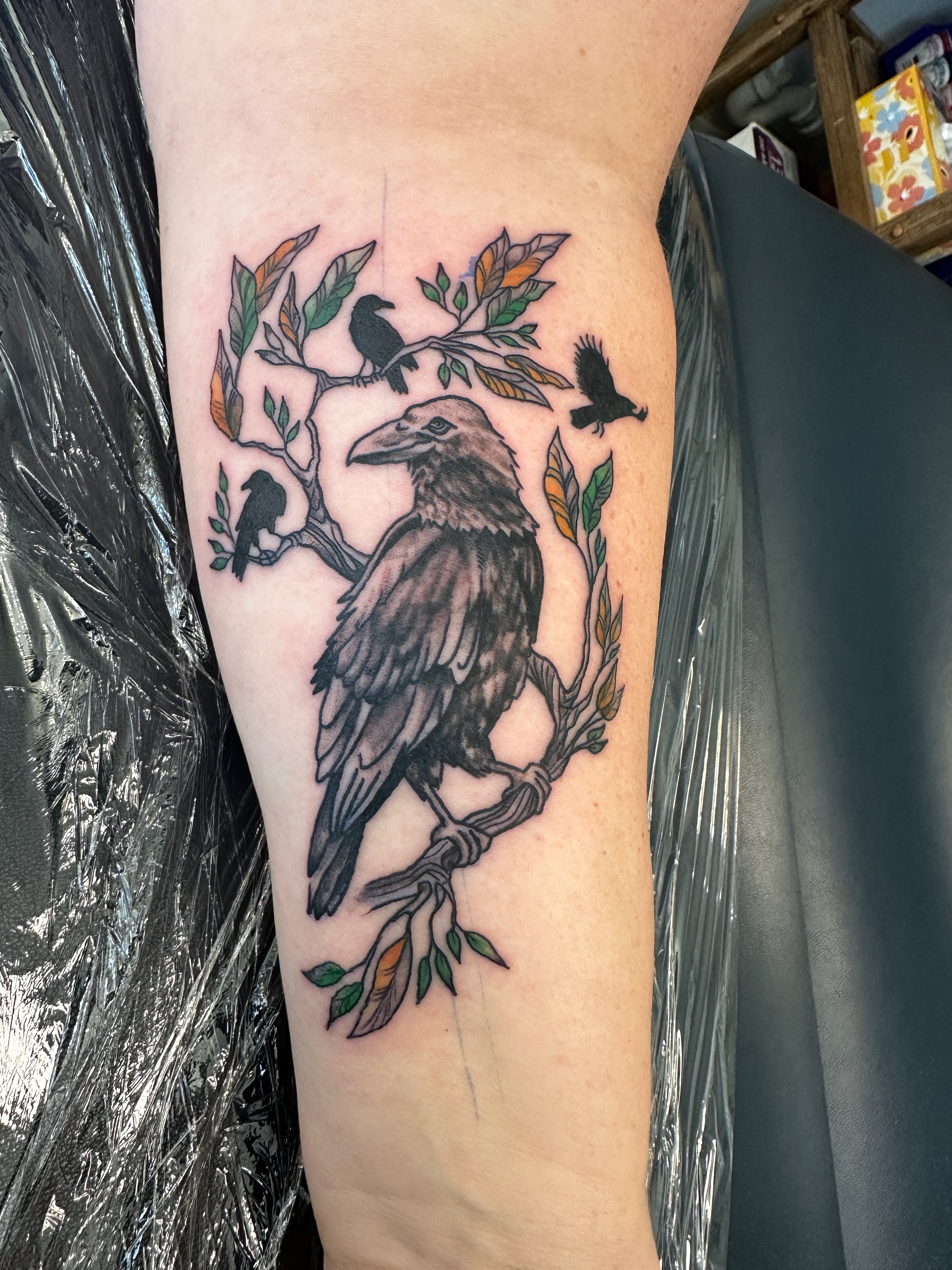 Tattoo of a large bird, likely an eagle, sitting on a tree branch with smaller birds perched and flying nearby, with green and orange leaves. Tattoo designed by Robin Reece