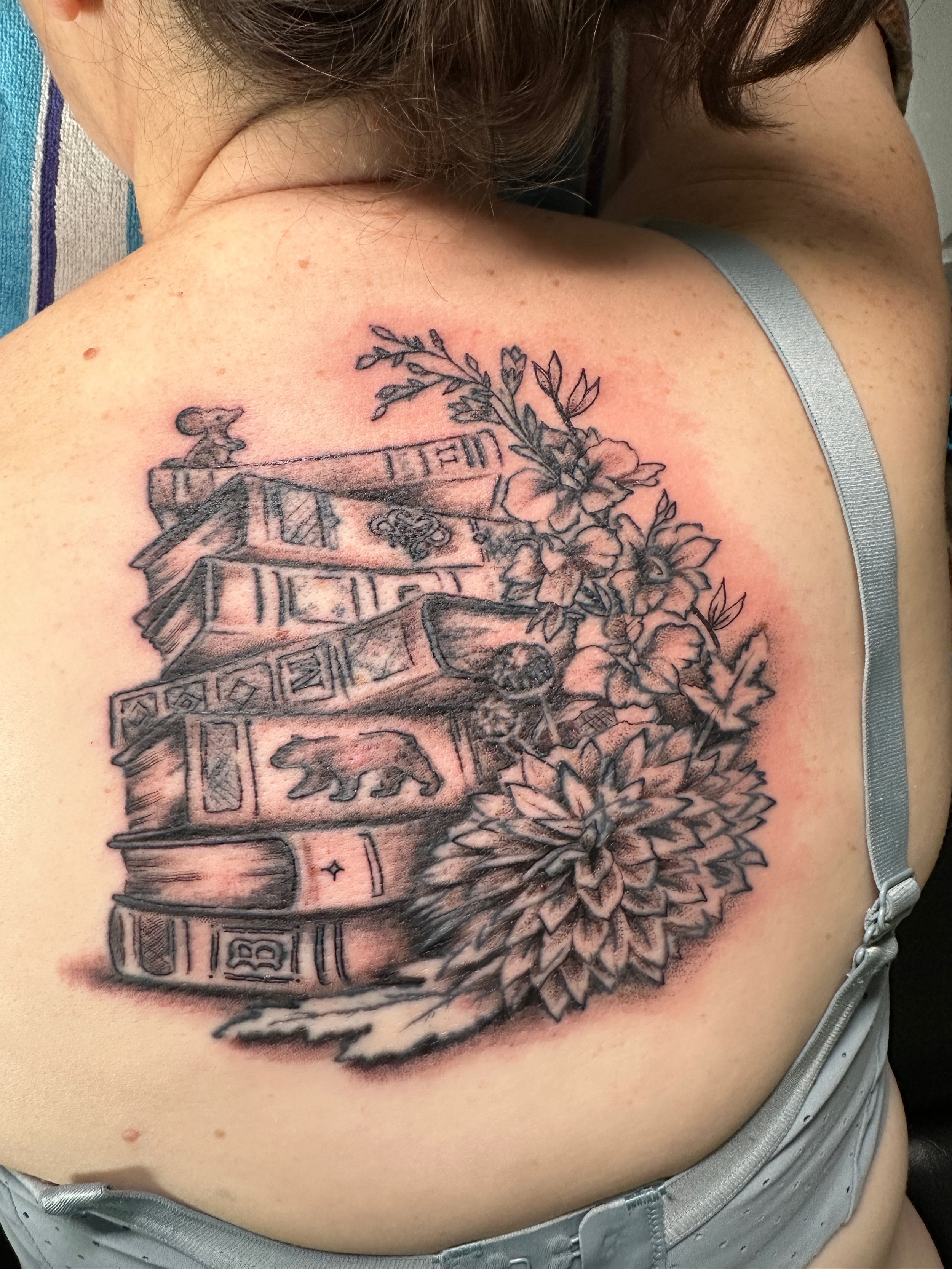 A shoulder tattoo featuring a stack of books with intricate details, surrounded by flowers and leaves, in black and gray ink. Tattoo designed by Robin Reece