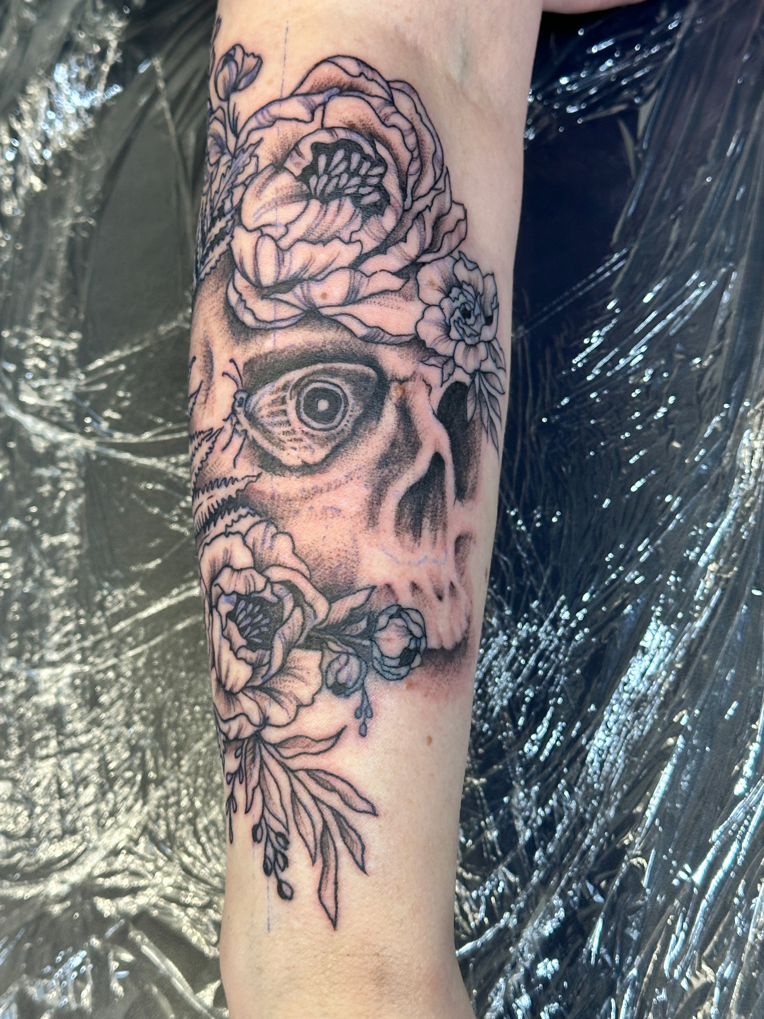 Black and gray tattoo of a human skull surrounded by flowers on a person's arm. Tattoo designed by Robin Reece