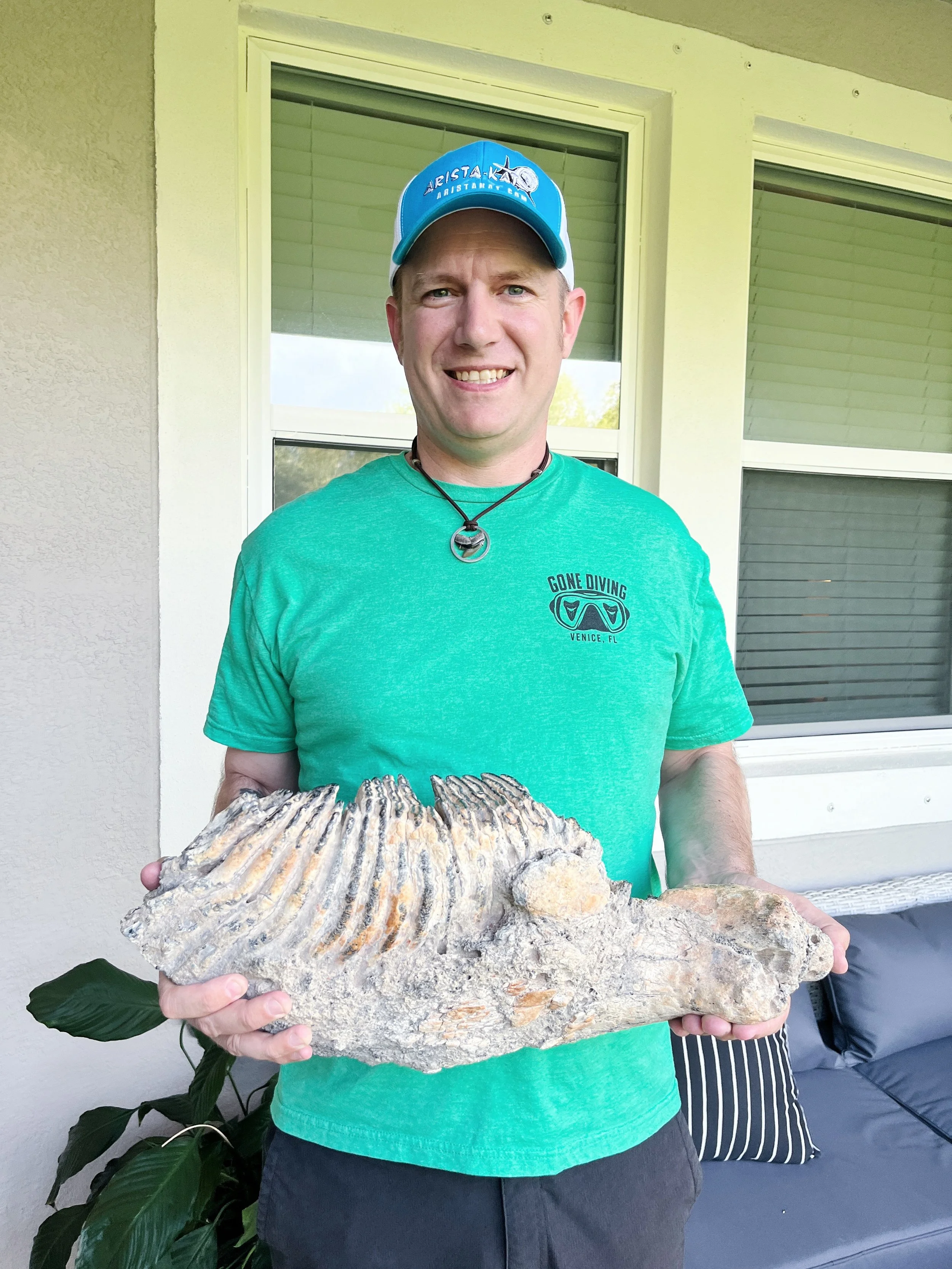 Man holding a large mammoth tooth jaw.  He is wearing a blue cap, a green T-shirt with a gone diving logo, and a necklace.