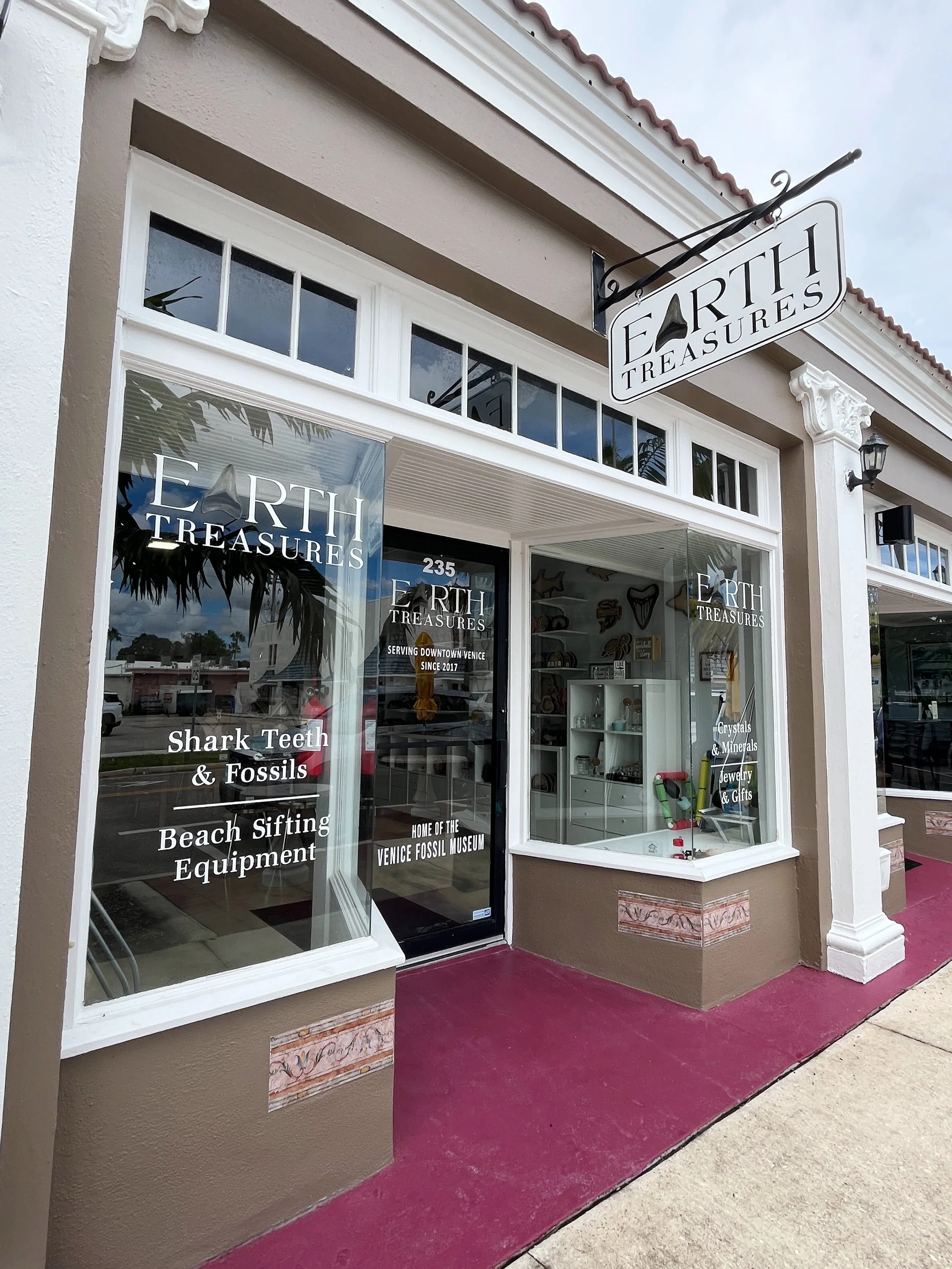 The storefront of Earth Treasures, a shop selling fossils, minerals, jewelry, and gifts, with large windows and a sign hanging above the entrance. The windows display the shop's name and items inside.