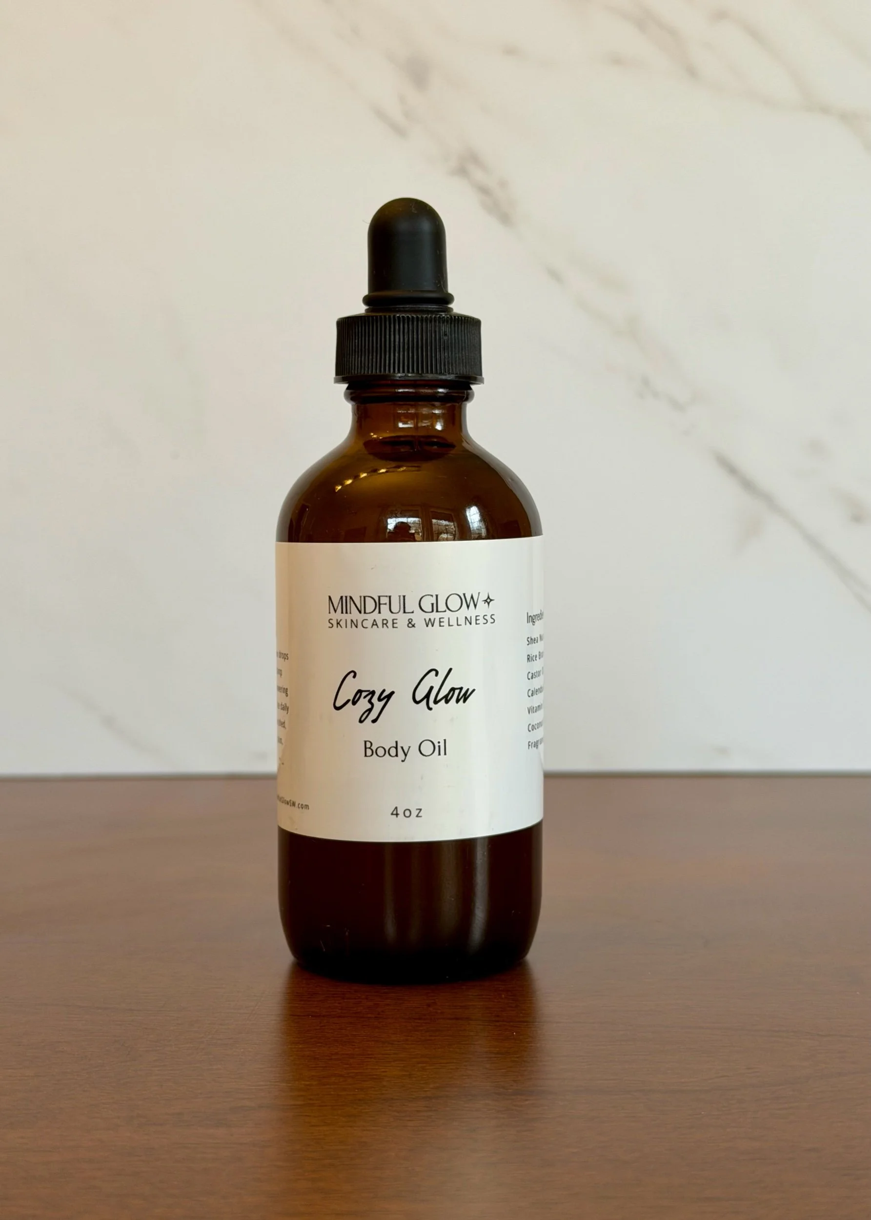 A brown glass bottle with a black dropper cap labeled 'Mindful Glow Skincare & Wellness, Cozy Glow Body Oil, 4 oz', sitting on a wooden surface with a white marble background.