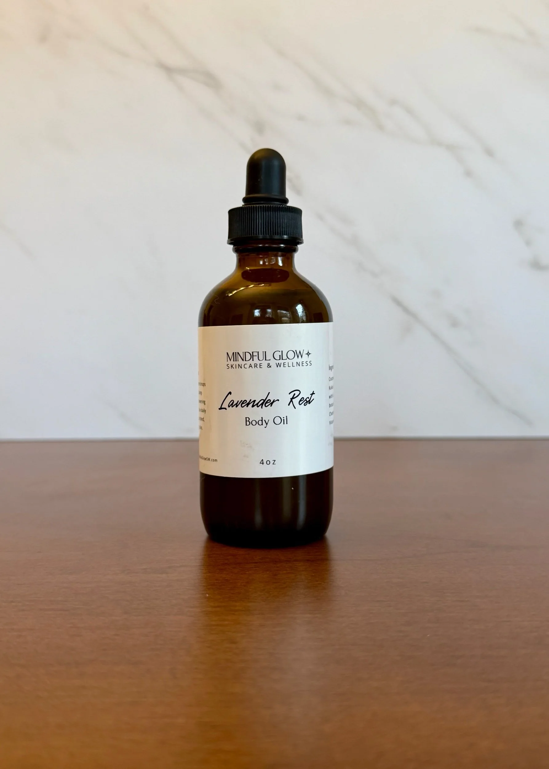 A small amber glass dropper bottle labeled 'Lavender Rest Body Oil' from Mindful Glow, placed on a wooden surface against a white marble background.