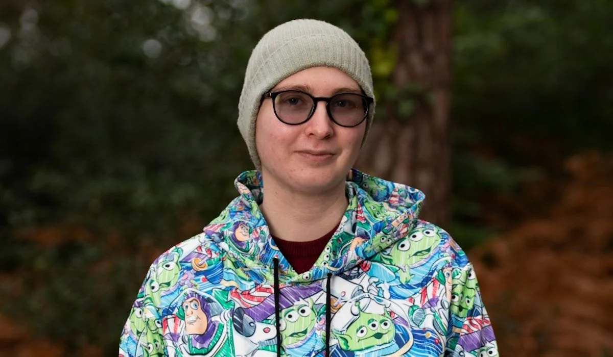 A close up image of the author, Ronald Barr, in the forest. He is wearing a brightly coloured hoodie, tinted glasses, and a beanie. On the surface, he appears to be happy.