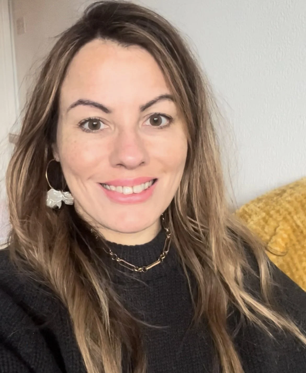 A smiling woman with long, wavy brown hair, wearing hoop earrings, a chain necklace, and a black top. She is sitting on a yellow textured couch and the background shows a plain wall.