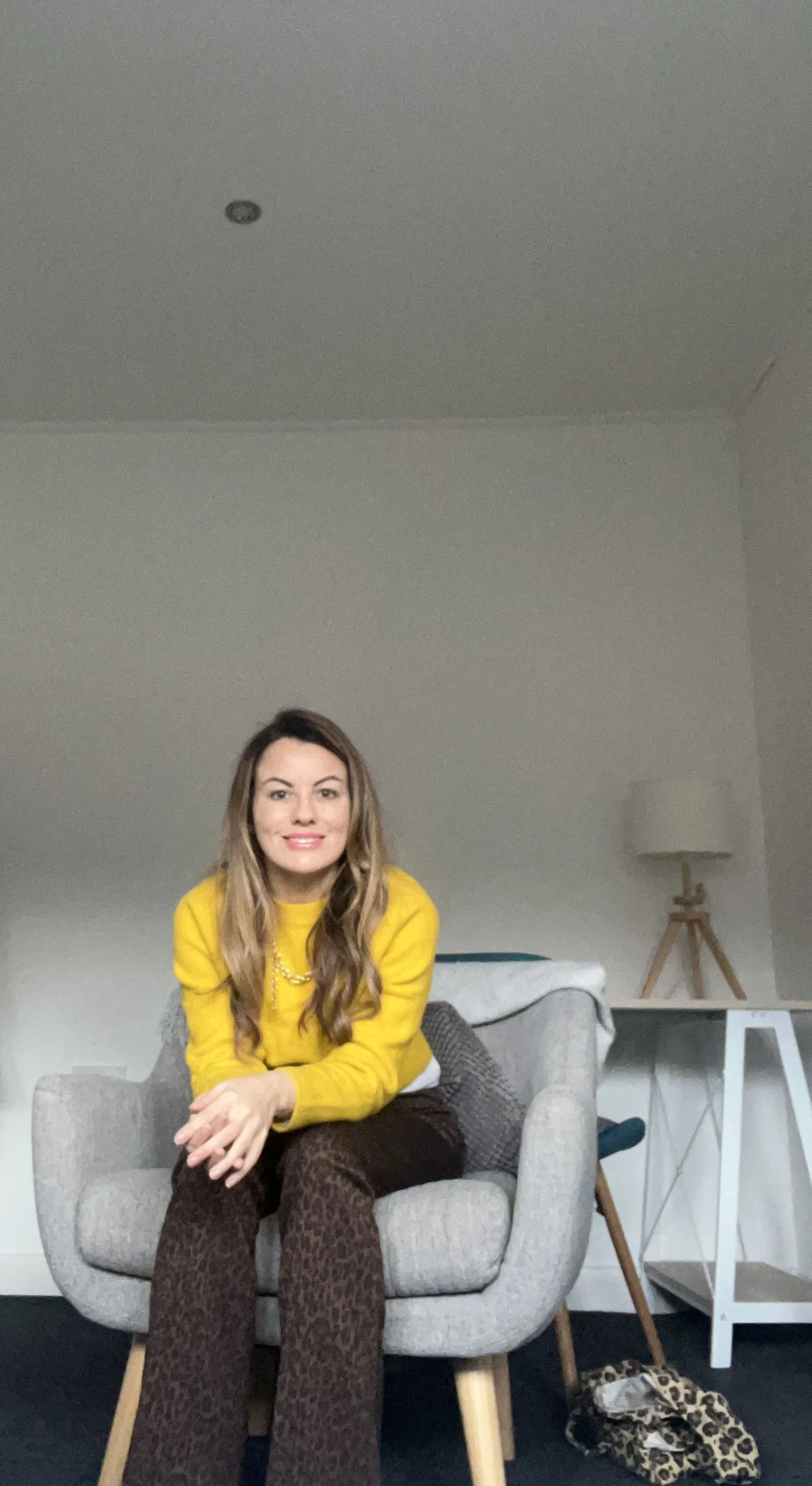 A woman with long, wavy hair wearing a bright yellow sweater and brown patterned pants is sitting on a light gray armchair in a room with minimalist decor, including a white side table, a tripod floor lamp, and a leopard print bag on the floor.