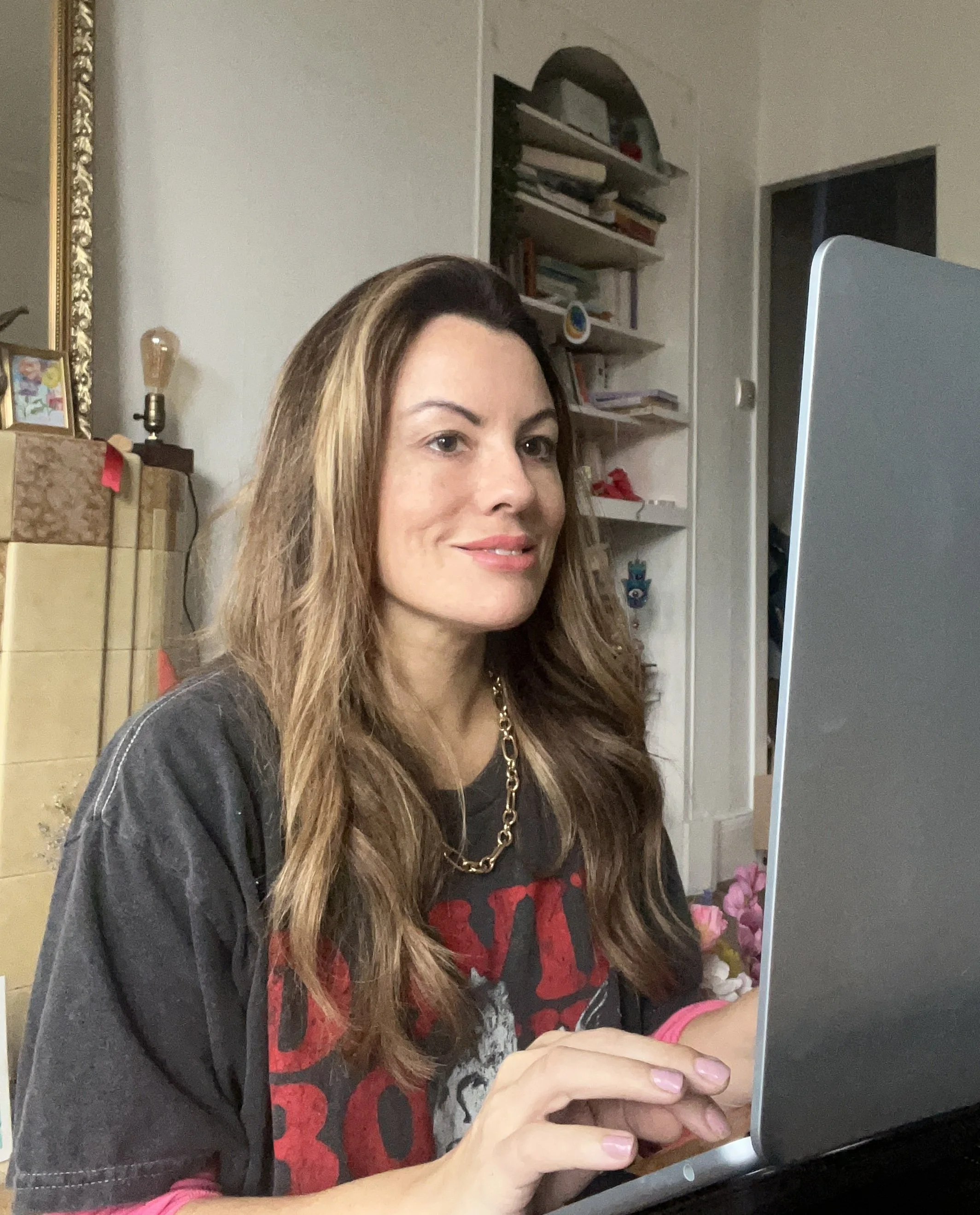 A woman with long brown hair working on a laptop in a room with bookshelves and decorations.
