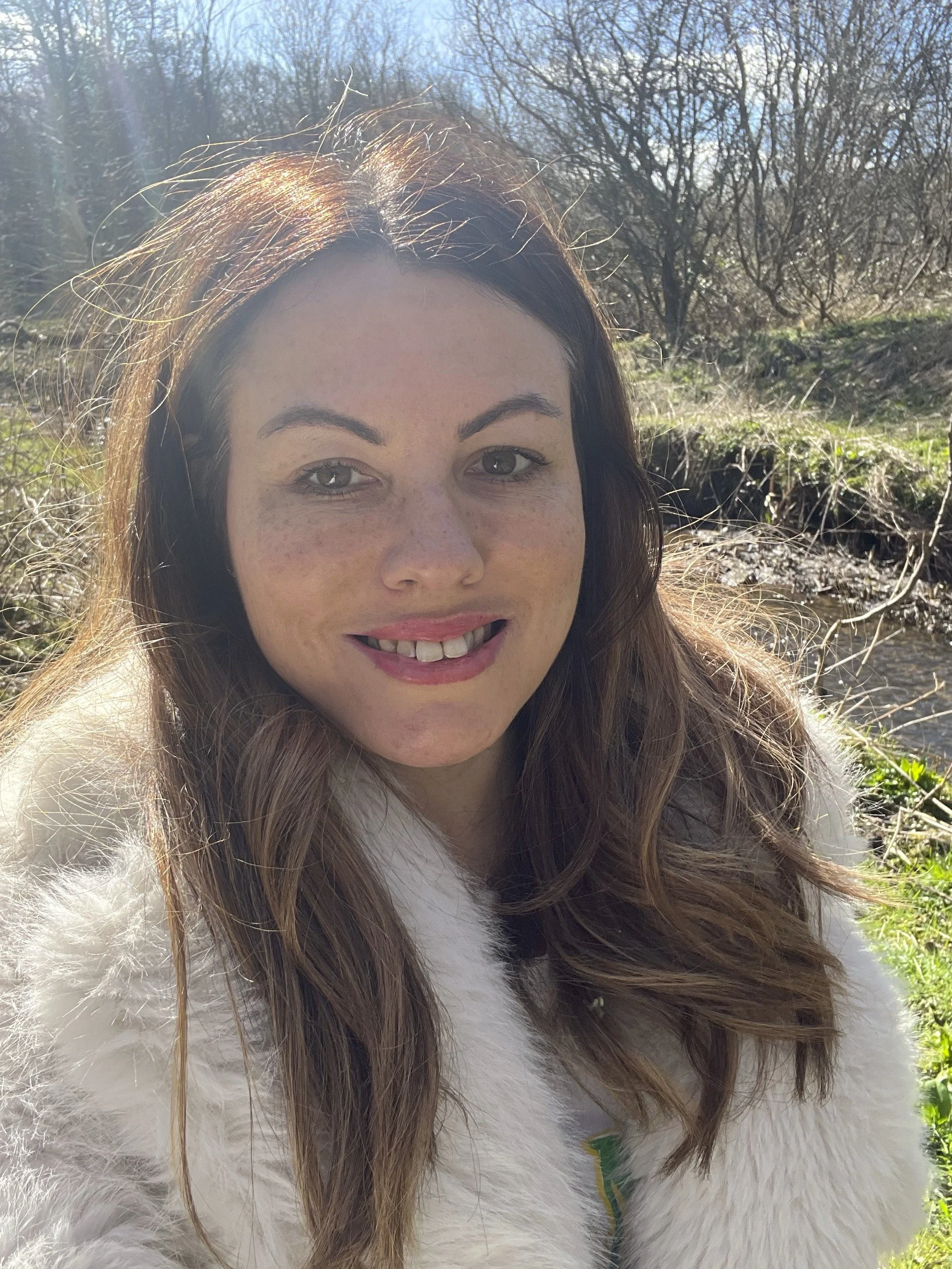 A woman with long brown hair, fair skin, and blue eyes taking a selfie outdoors in sunlight, wearing a white fuzzy coat with nature in the background.