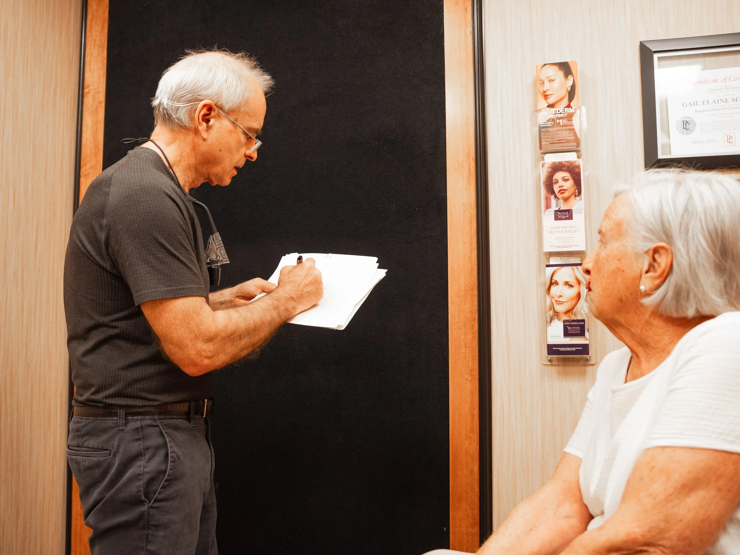 Dr. Chariker taking notes while talking to a woman seated in a treatment room, with promotional materials and a certificate on the wall in the background.