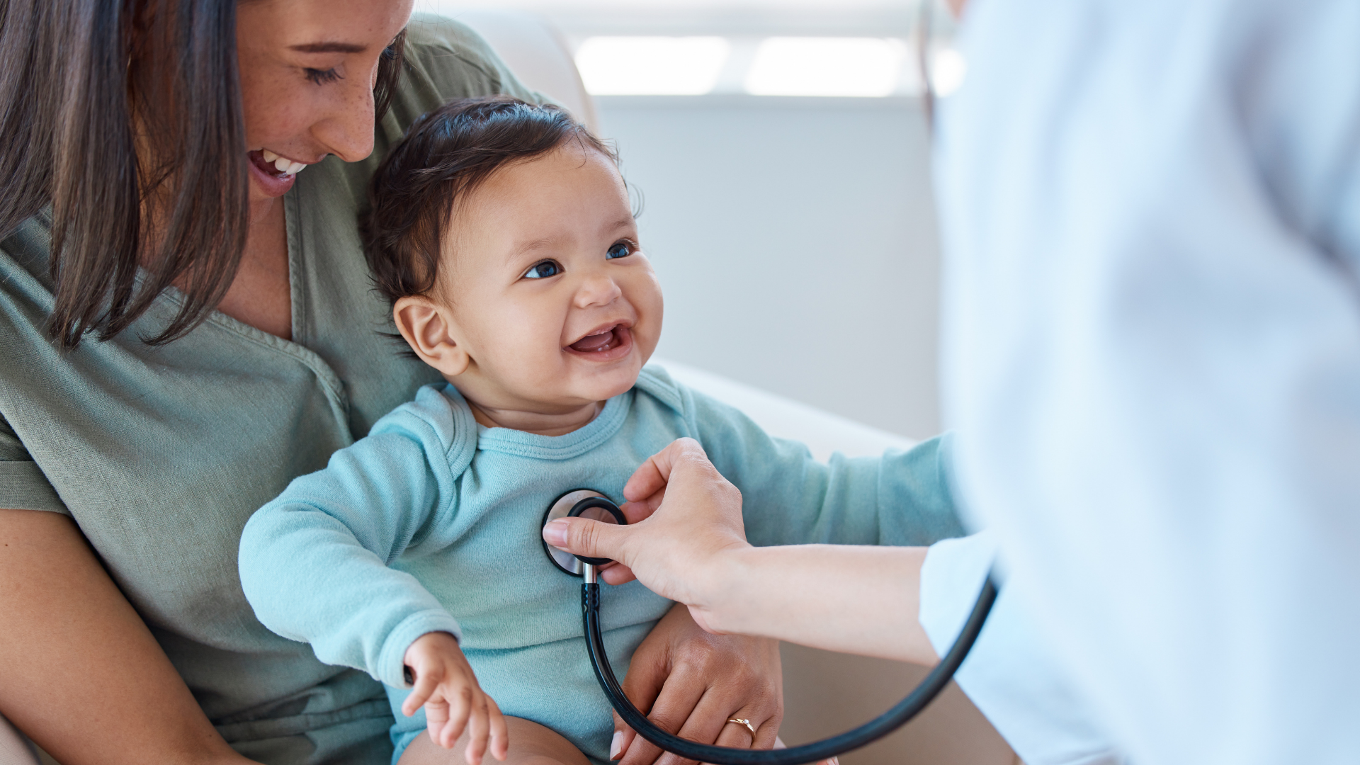 A smiling baby wearing a blue shirt is sitting on a woman's lap as a healthcare professional uses a stethoscope to listen to the baby's chest. The woman, likely the mother, is looking at the baby lovingly. The setting appears to be a medical exam room with bright lighting.