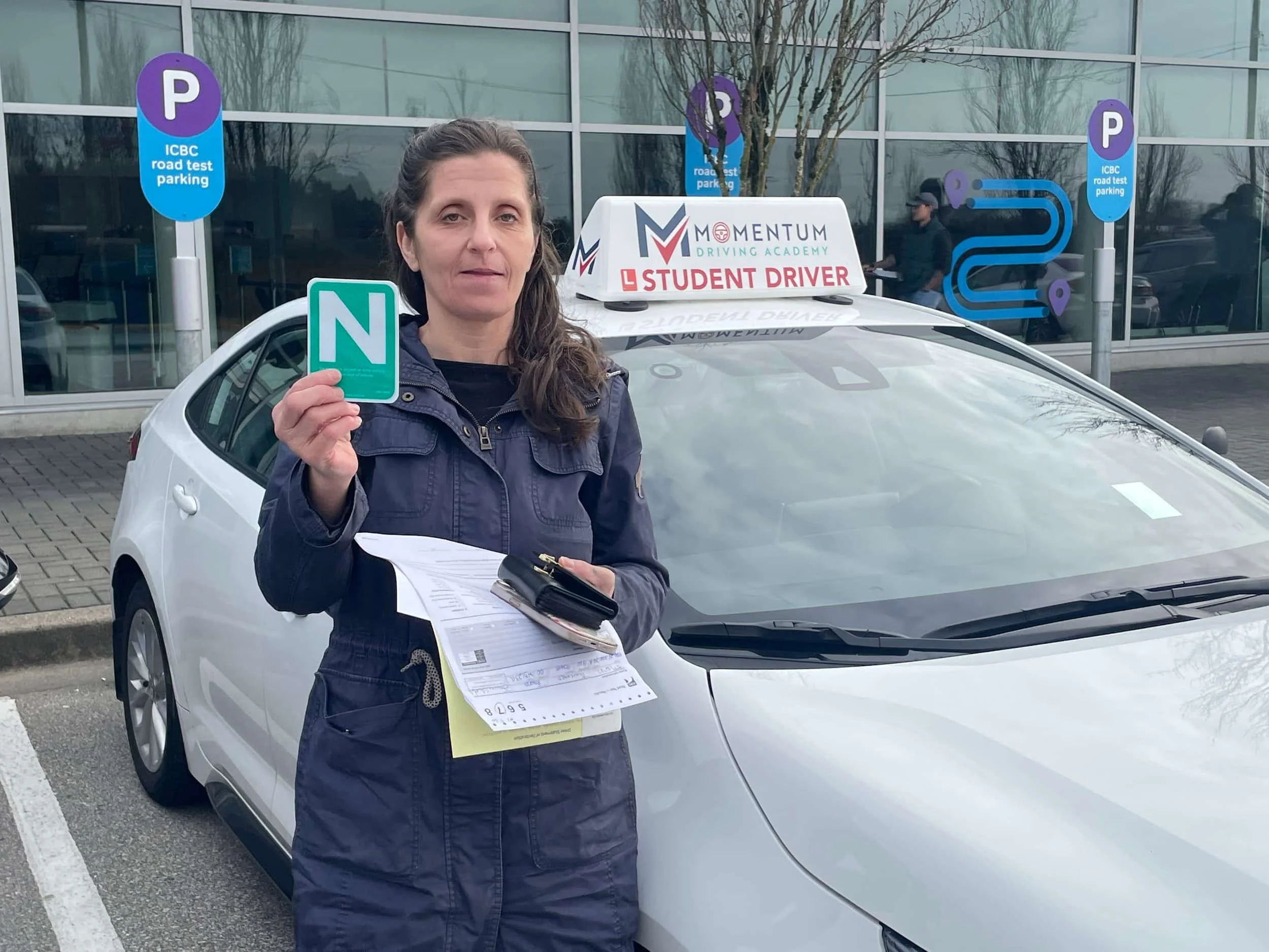 Woman holding a green N sign beside a white student driver car in a parking lot, holding papers and a wallet.