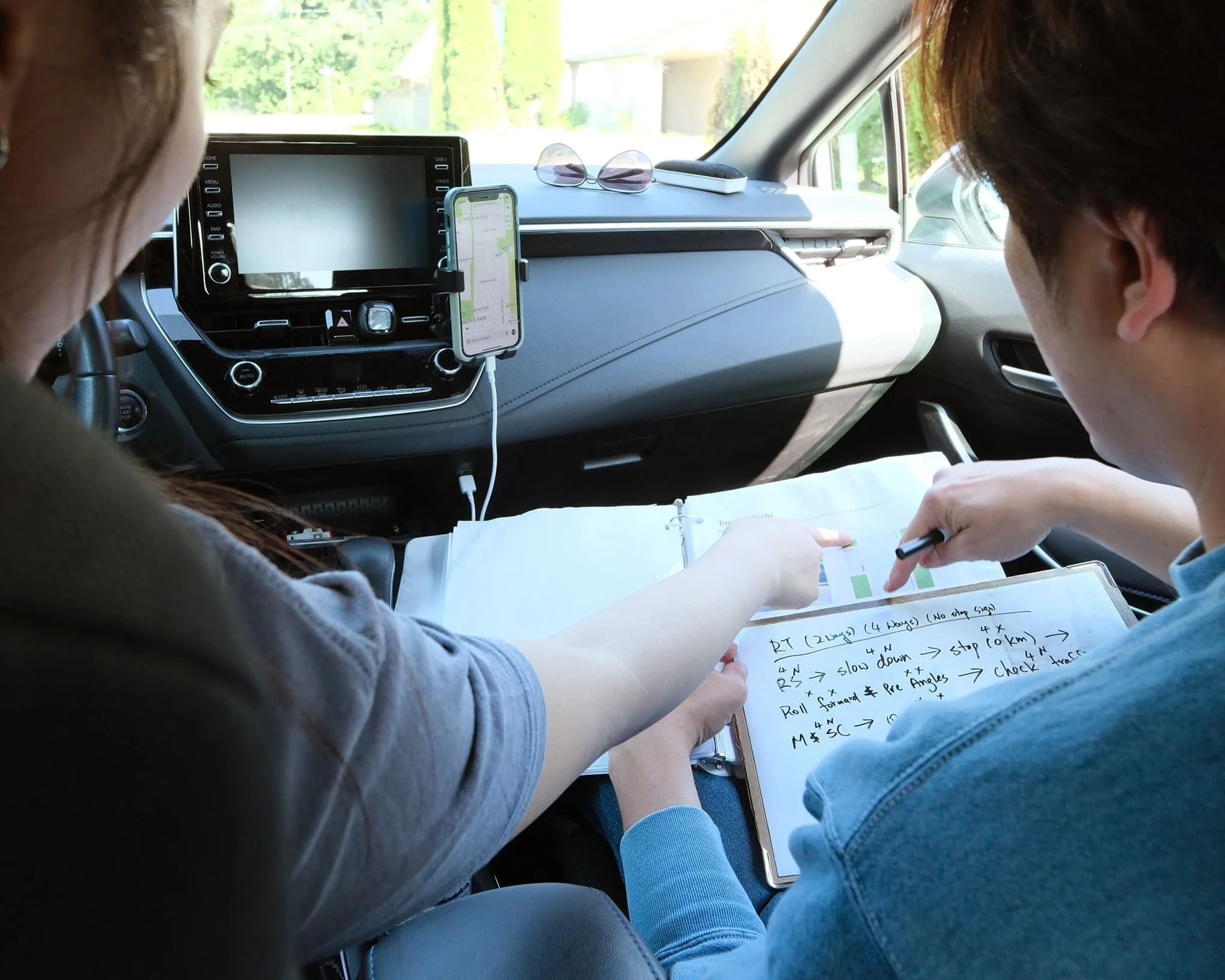 Two people sit in a car; one points at notes in a binder while the other writes on a whiteboard with markers.