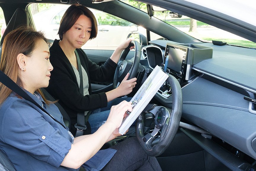 Two women sit in a car; a female instructor reviews paperwork on a clipboard while the other grips the steering wheel, listening attentively.