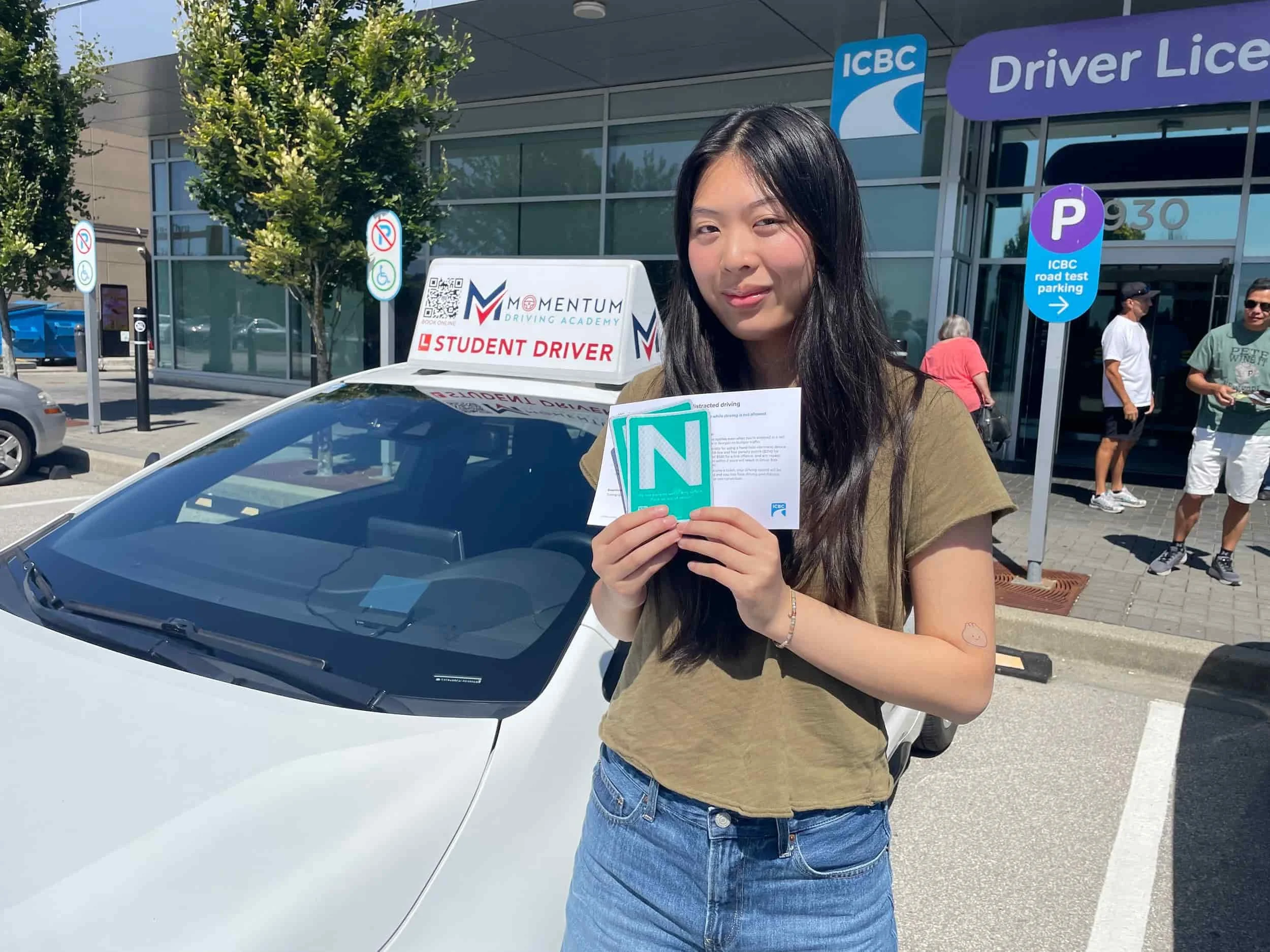 Young woman holding a learner’s license and papers stands by a car with a “Student Driver” sign outside a driver licensing office.