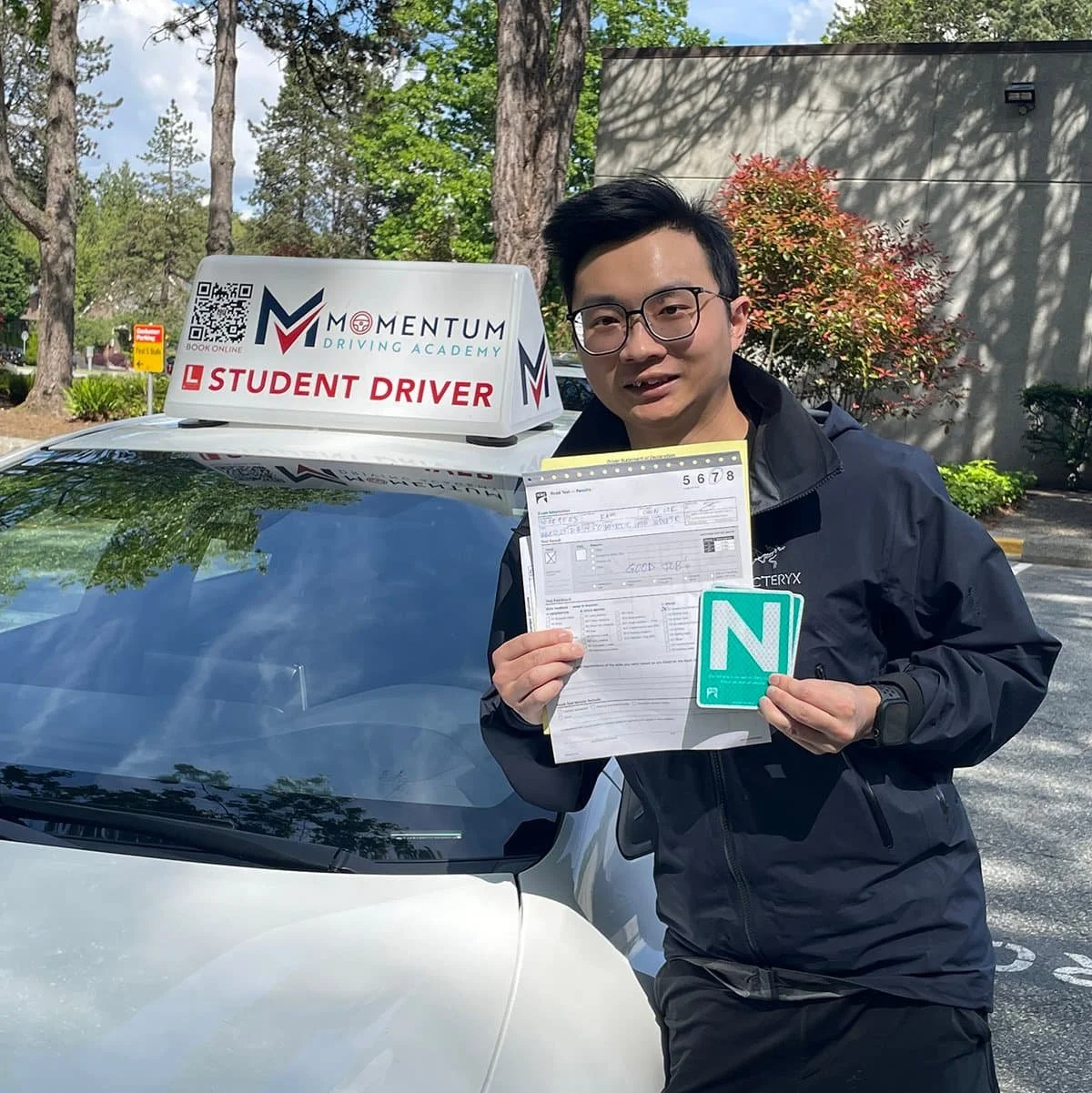 A person stands by a car with a Student Driver sign, holding paperwork and a green N for novice driver.