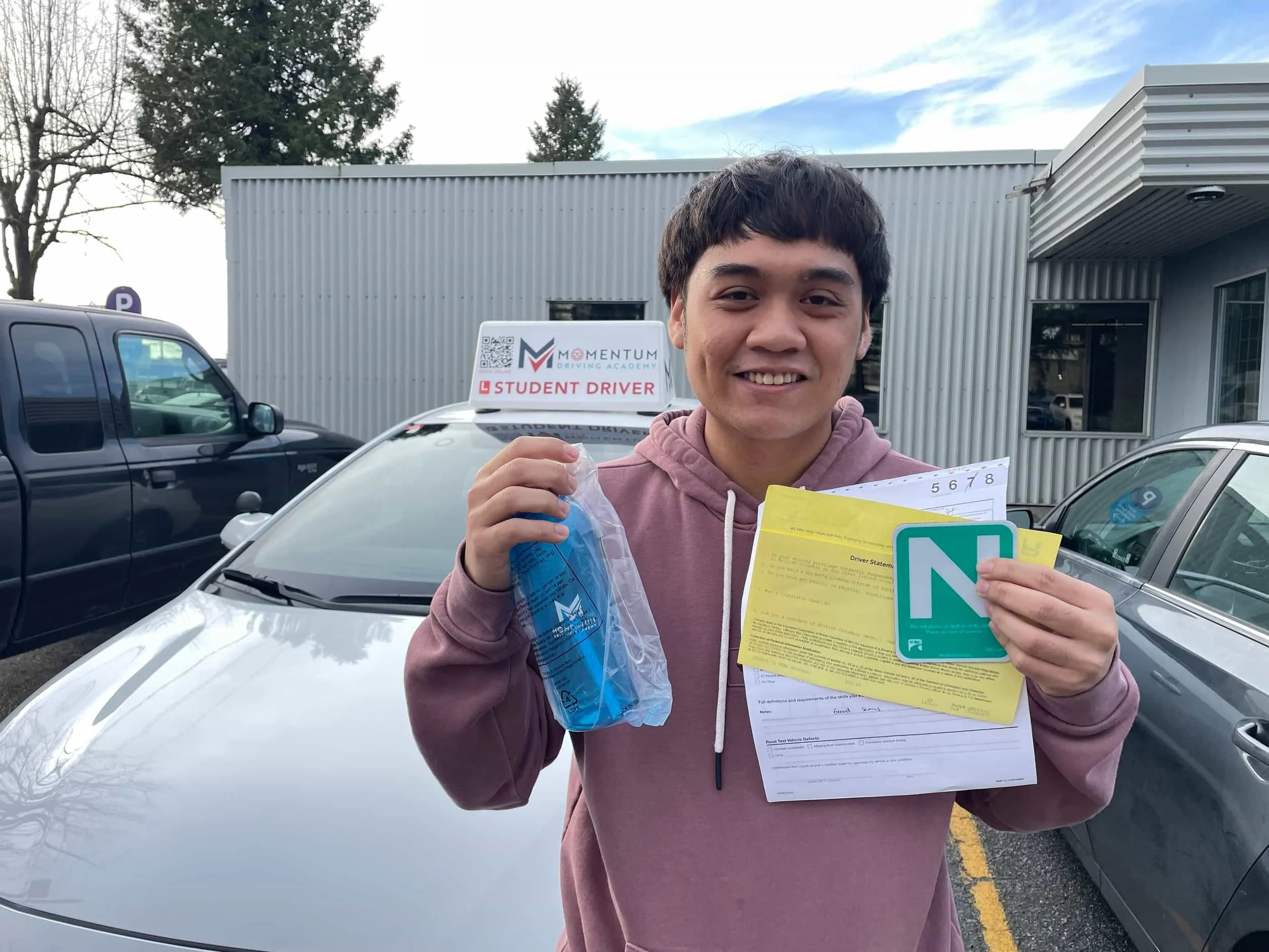 Smiling person in pink hoodie holds paperwork and a green N sign in front of a car with a Student Driver sign on top.