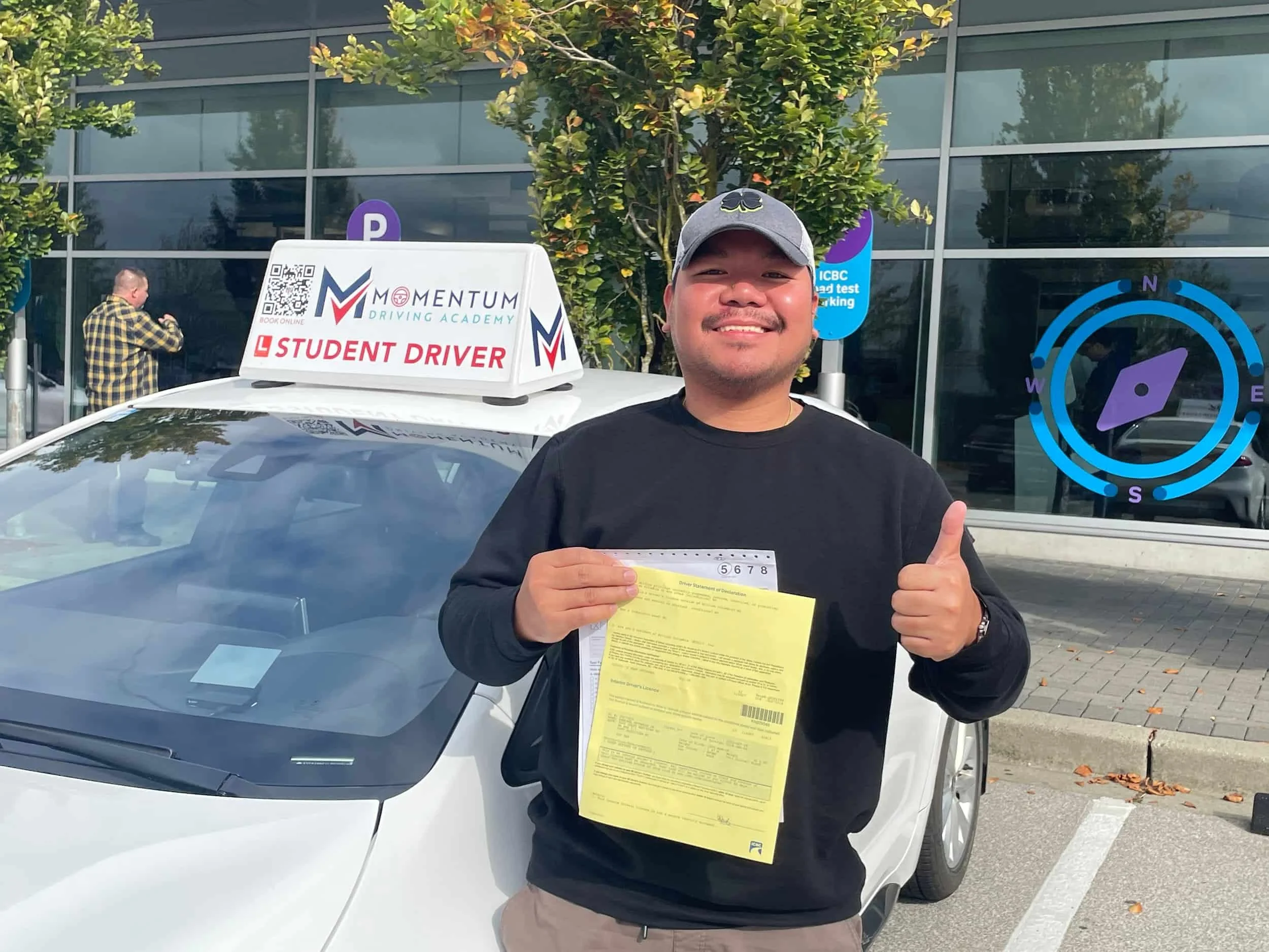 A smiling person stands by a car with a Student Driver sign, holding papers and giving a thumbs up.
