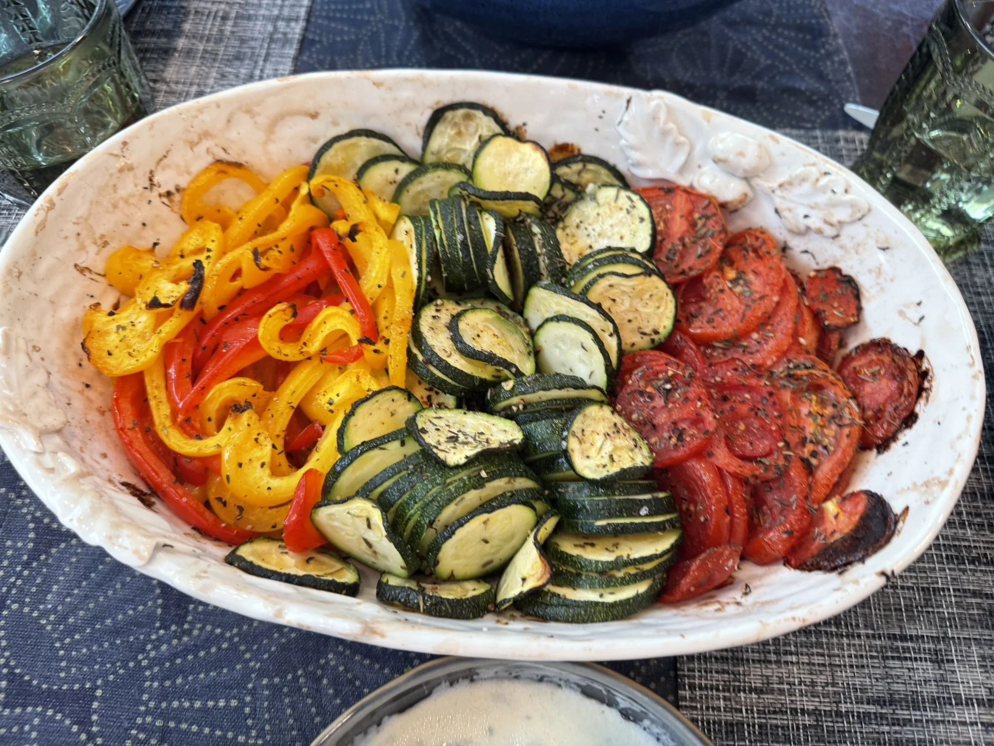 A white bowl filled with baked vegetables including sliced zucchini, red and yellow bell peppers, and tomatoes, on a dark woven placemat.