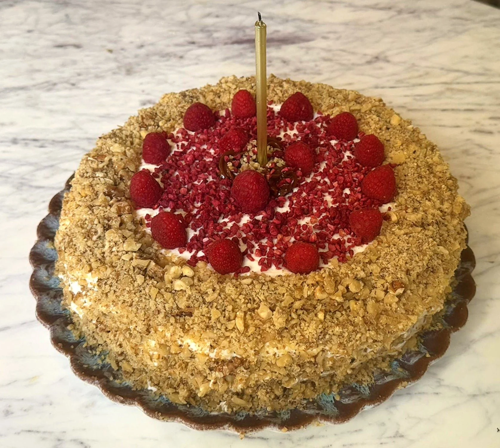 Round birthday cake with crushed nuts on the sides, topped with raspberries, crushed red topping, and a single white candle in the center, on a marble surface.