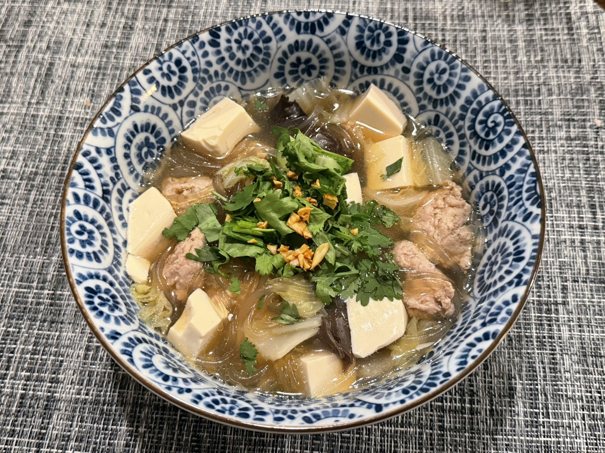 A bowl of Thai glass noodle soup with cubes of tofu, greens, chopped garlic, and herbs on top, served in a decorative blue and white bowl on a textured surface.
