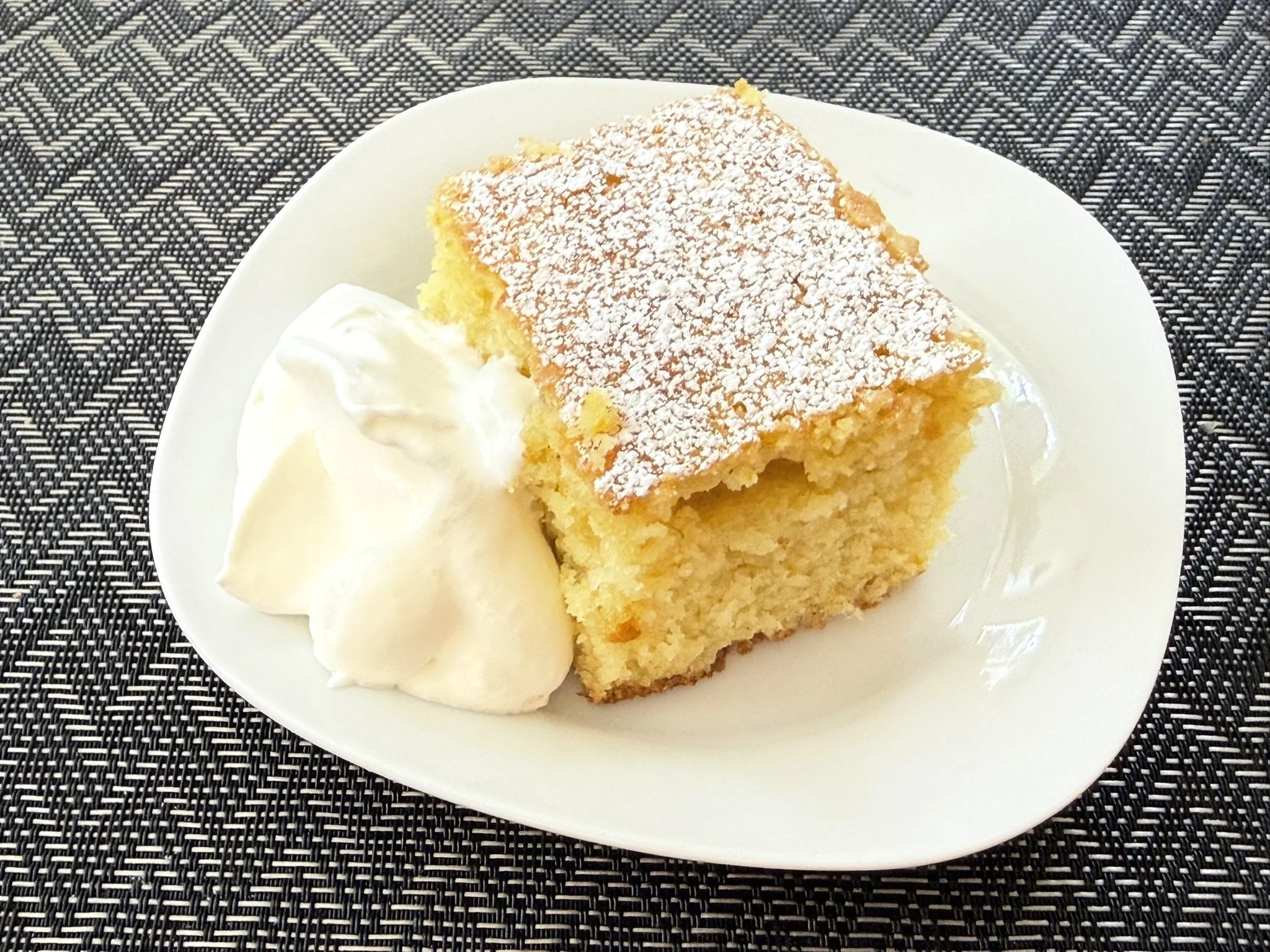 A white plate holding a slice of olive oil cake dusted with powdered sugar and a dollop of whipped cream, set on a black and white woven placemat.