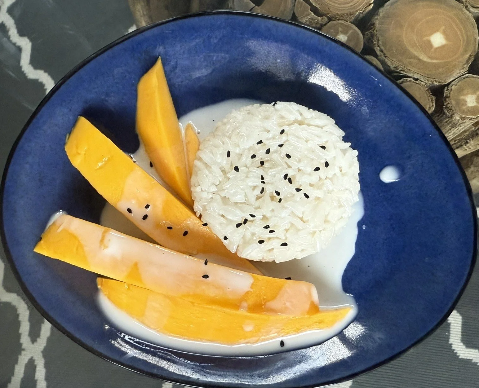 A bowl of Thai sticky mango rice dessert topped with black sesame seeds, in a blue bowl with a white coconut milk sauce.