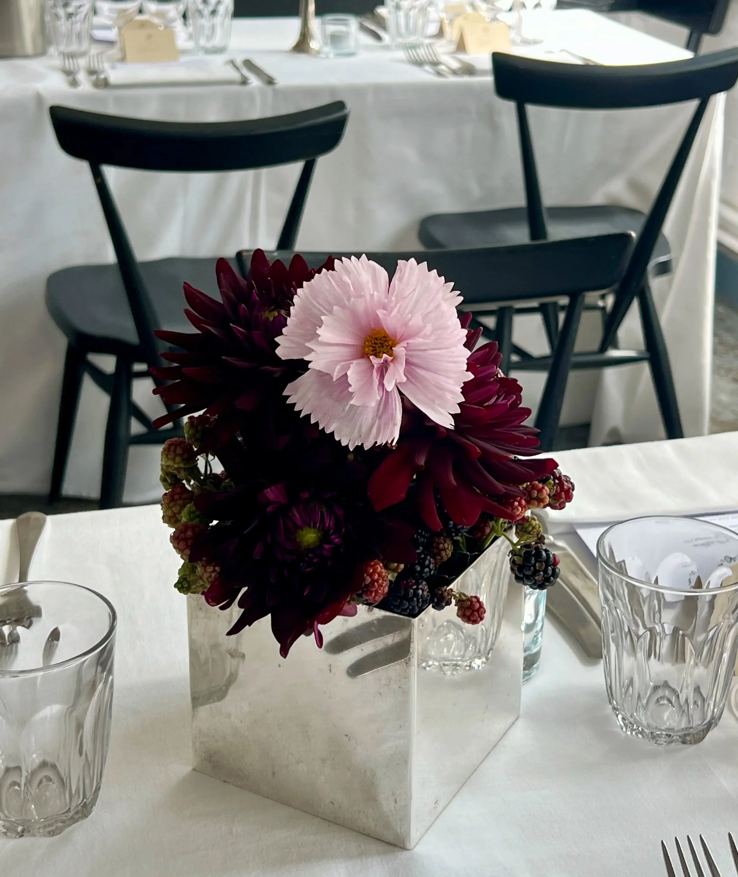 A floral centerpiece with pink, dark red, and blackberries in a square white marble vase, surrounded by empty clear glass drinking glasses, tableware, and silverware on a white tablecloth.