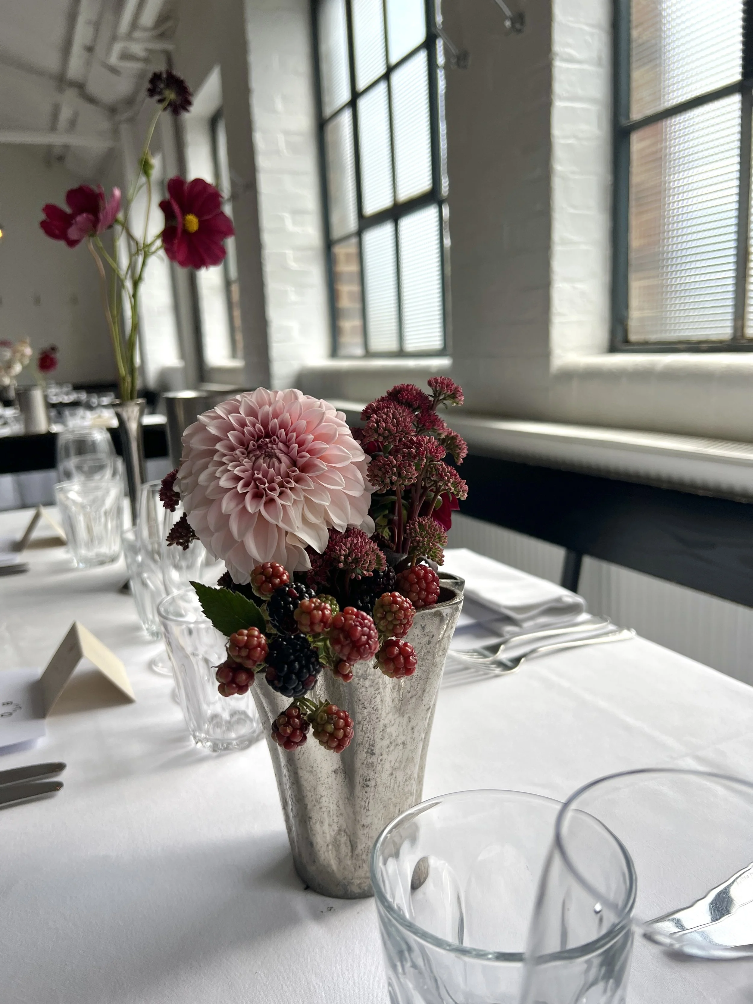 A table with a vase of pink, white, and purple flowers and berries, set with glassware, in a room with large windows and white brick walls.