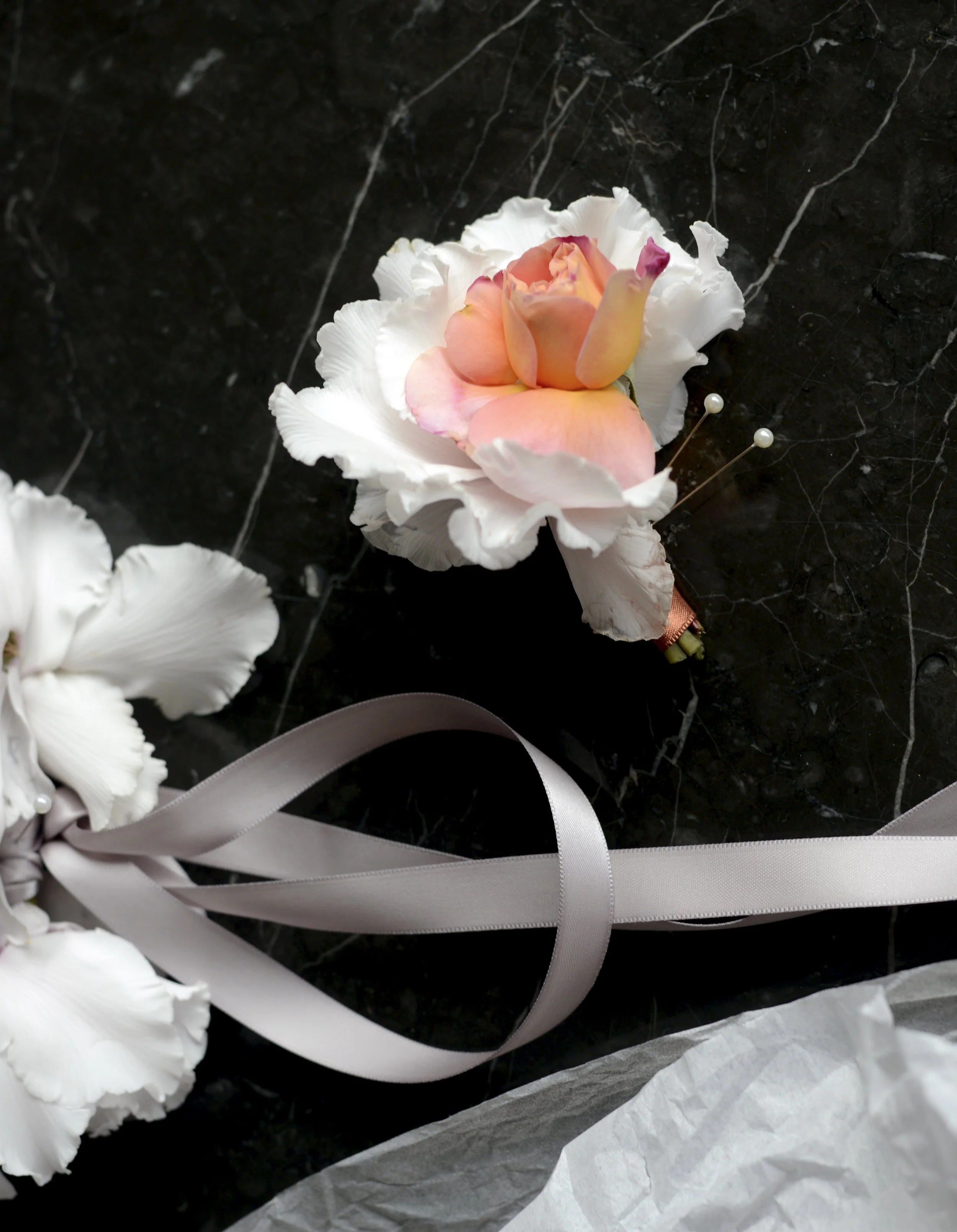 Close-up of a pink and white artificial flower boutonniere on black marble surface, with grey satin ribbon and white tissue paper.