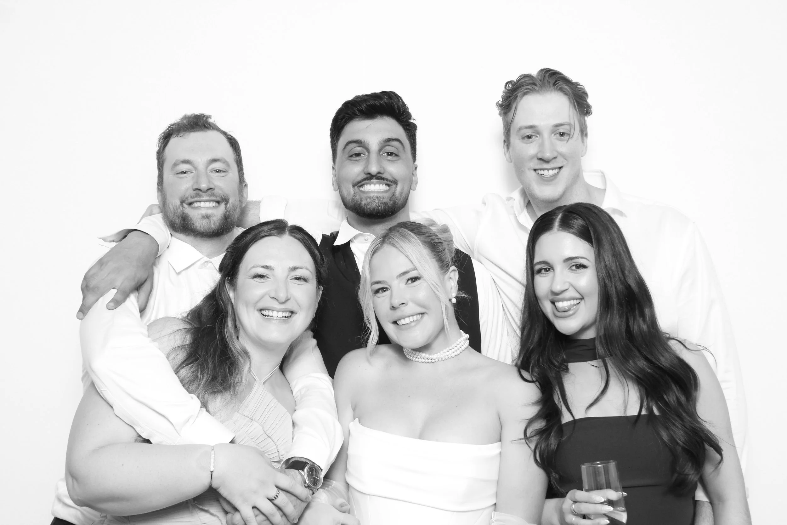 Group of six friends smiling and posing together at a celebration, with some holding drinks, in black and white.