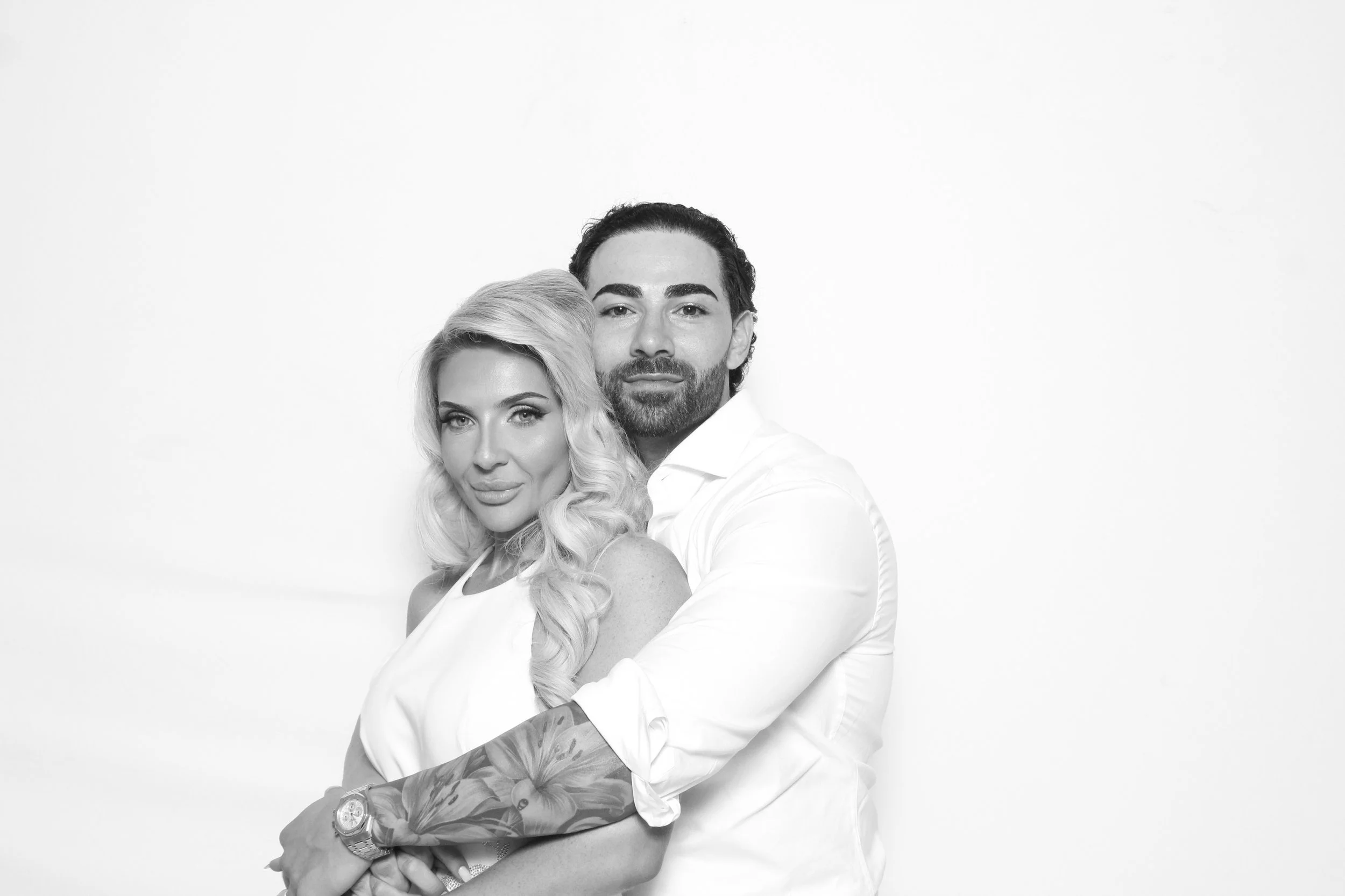 A black and white photo of a man and a woman standing close together, with the woman in front and the man slightly behind. The woman has long, wavy blonde hair and is looking directly at the camera. The man has dark hair, a beard, and is also looking