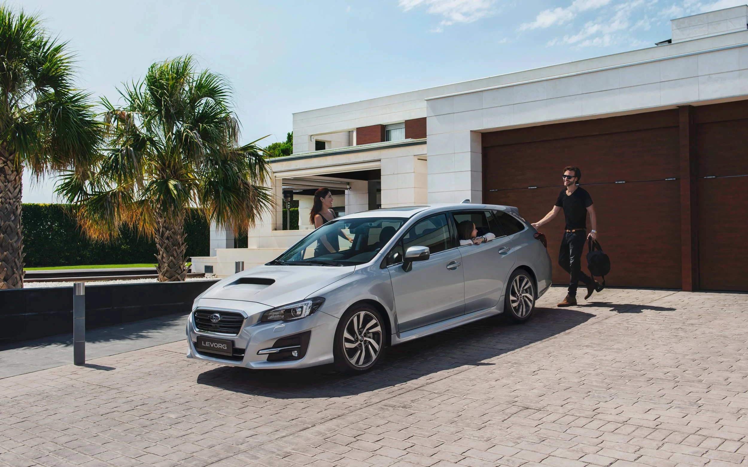 A silver station wagon parked in front of a modern house with palm trees. A woman is talking to a man with sunglasses, who is carrying a black bag, while a girl sits inside the car.