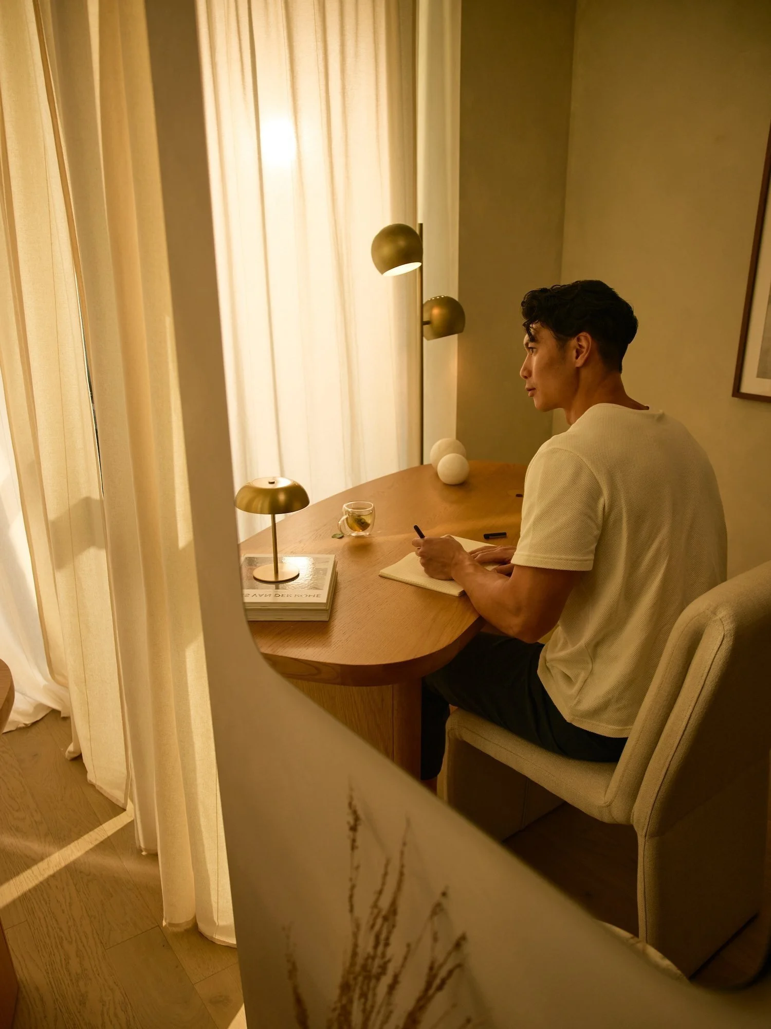 A man in a white t-shirt sitting at a wooden desk with a notepad, smartphone, and a glass of drink, seen through a circular cutout in a wall, with warm lighting and beige curtains in the background.