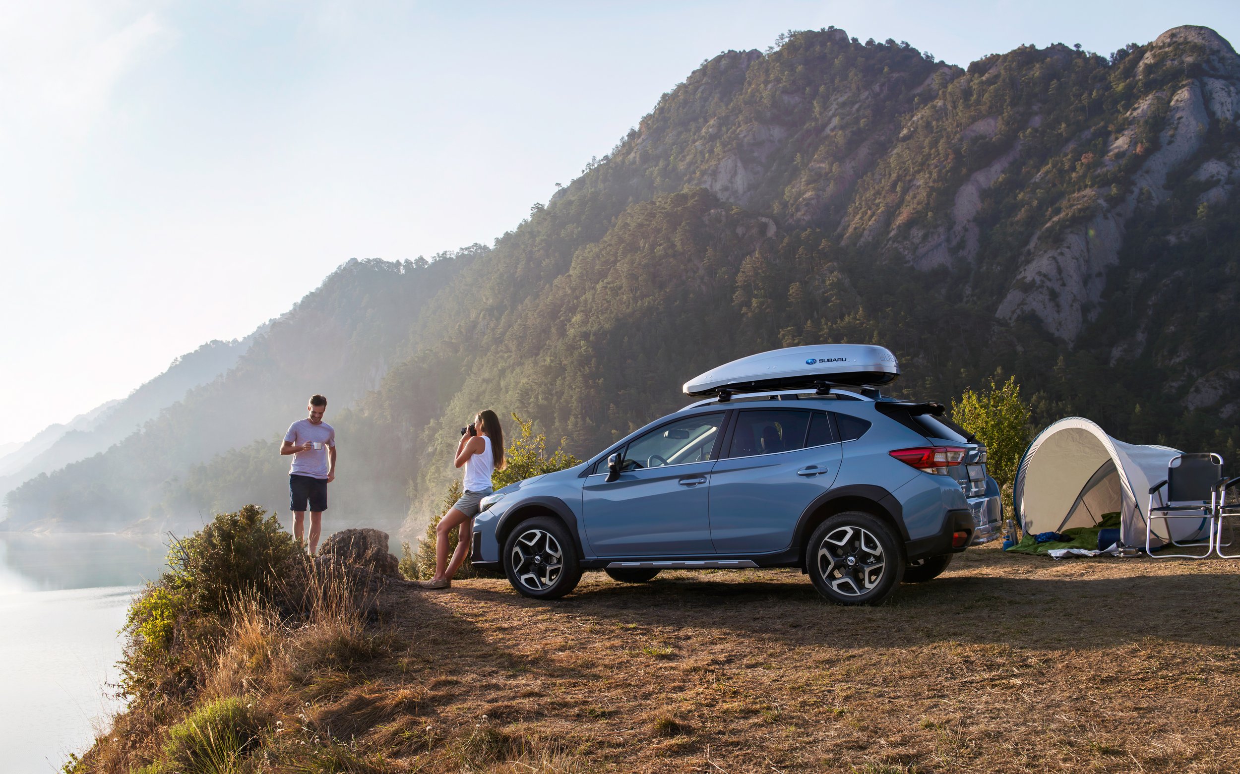 Two people camping by a lake with a mountain backdrop. A blue Subaru SUV with a roof cargo box is parked nearby, and a small tent and camping chair are set up on the grass.