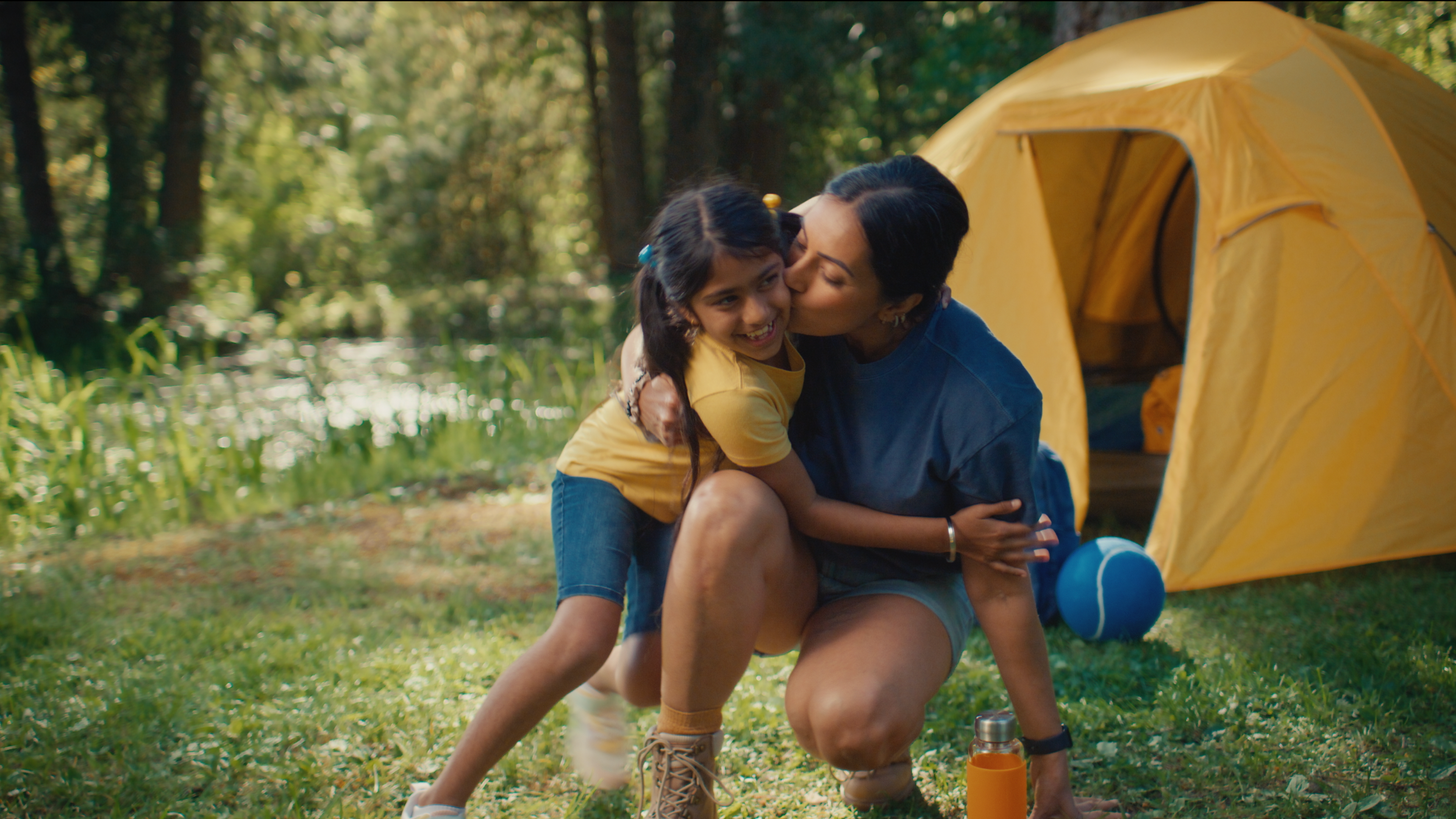 A woman and a young girl embrace and share a kiss in front of a yellow camping tent in a forested area, with trees and water in the background.