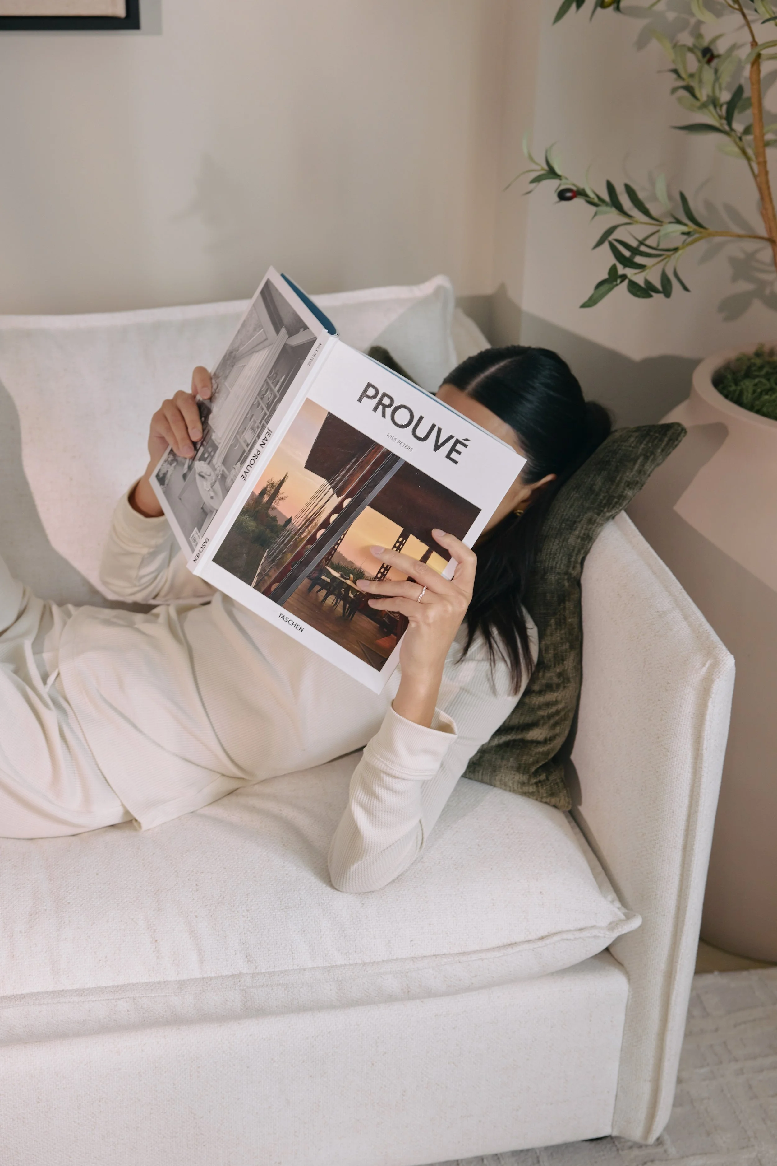 A woman lying on a beige sofa, reading a book titled 'PROUVÉ' with images of interior spaces, while resting her head on a dark green pillow and wearing a cream-colored outfit. There is a potted plant with green leaves in the background.