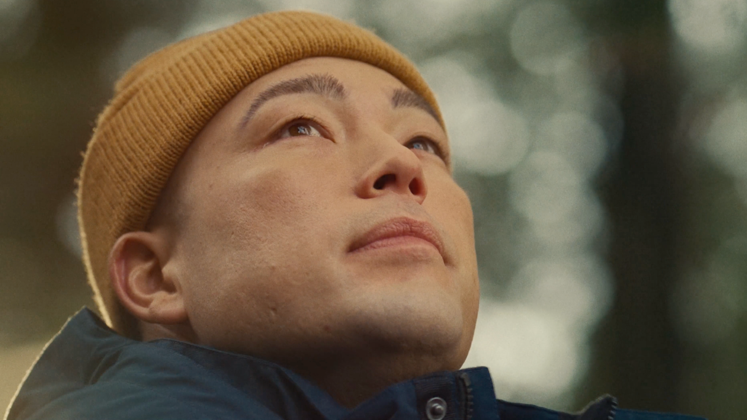 Close-up of a person wearing a yellow beanie and a dark jacket, looking upward with a serene expression against a blurred outdoor background.