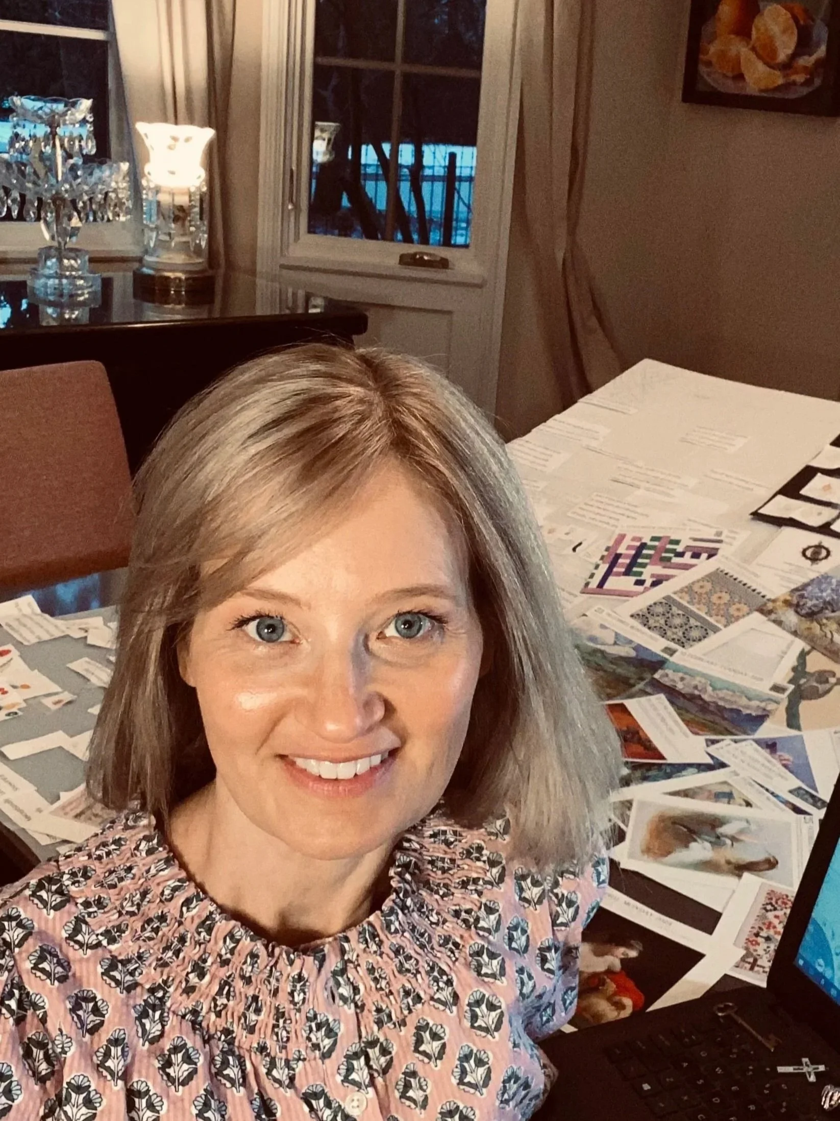 Seanra with shoulder-length blonde hair smiling at the camera, sitting at a table covered with papers, photographs, and printed materials, in a room with a window and a sideboard with crystal glasses and a lamp.