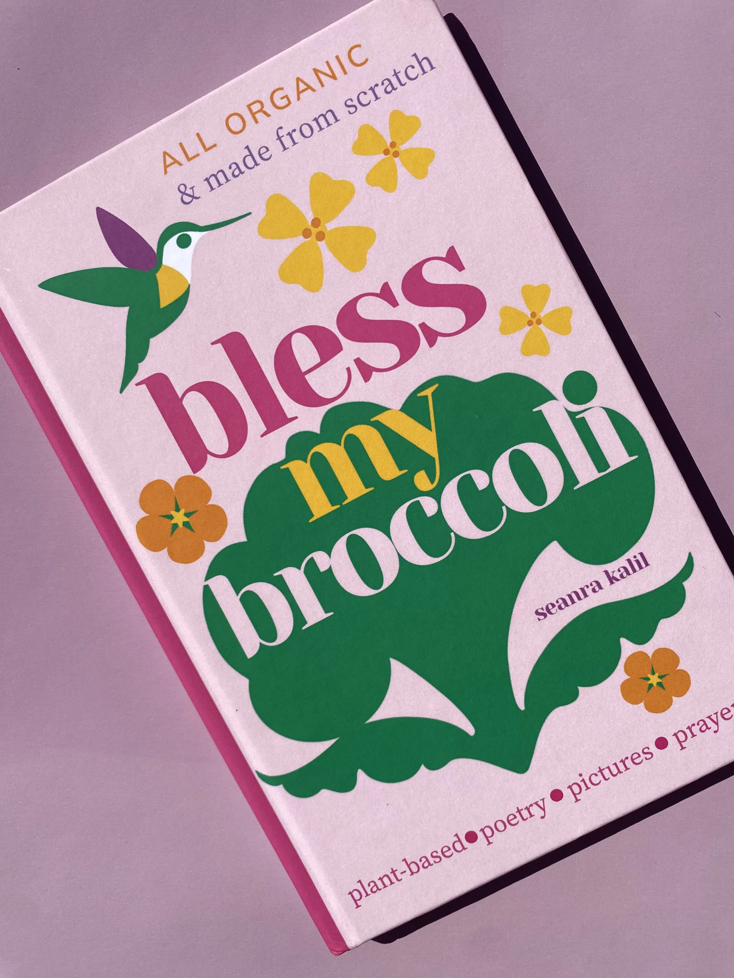 A book titled "Bless My Broccoli" by Seanra Kalil. The cover is pink with illustrations of a hummingbird, butterflies, and flowers. It mentions that the content is plant-based poetry, pictures, and prayers.