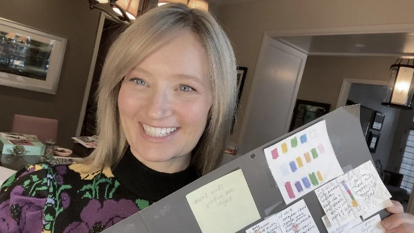 Seanra Askwith Kalil with blonde hair smiling and holding a gray board with colorful sticky notes and handwritten notes in a home setting.