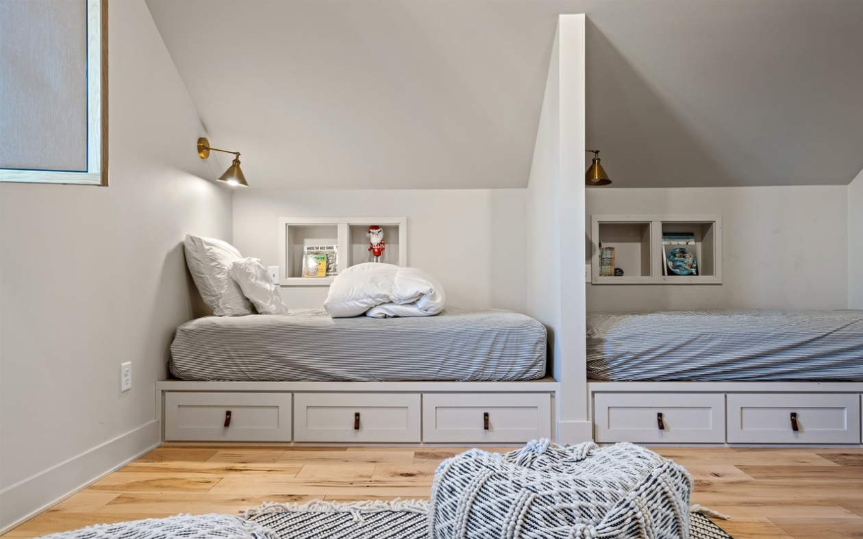 Two beds in a room with built-in shelves and wood drawer storage, separated by a partial wall, with wall-mounted lamps and light-colored walls and floors.
