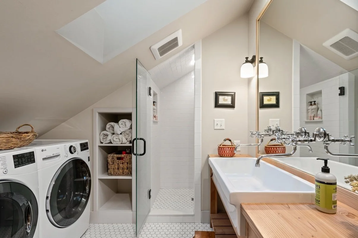 Bathroom with washer and dryer, a glass shower stall, a large mirror, a white sink with a wooden countertop, towels inside a shelf, framed pictures, and toiletries.