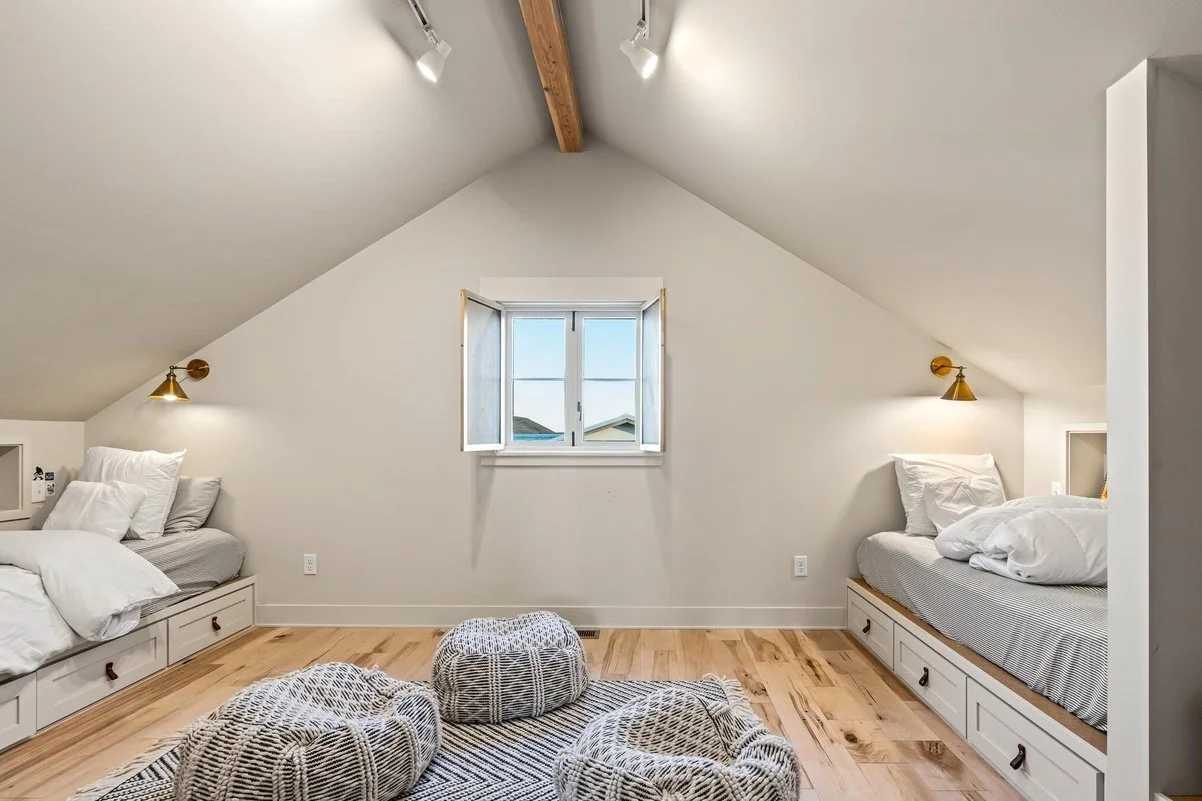 A bedroom with two twin beds built into the walls, each with white bedding and gold wall-mounted reading lamps. There is a small window in the center of the back wall, and a wooden beam runs along the ceiling. The room has light wood flooring and thr