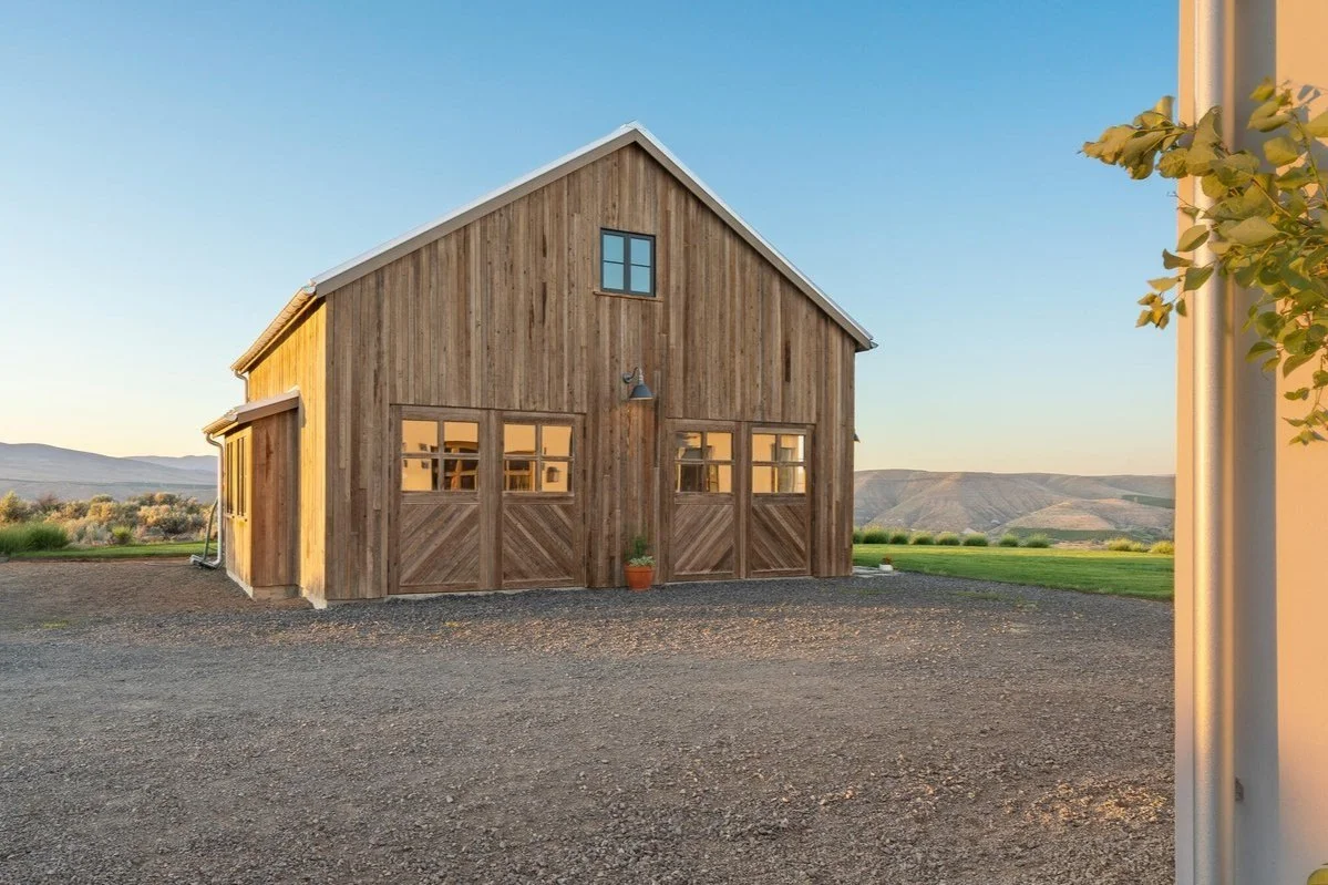 A wooden barn with sliding garage doors and small windows, situated on a gravel lot with a grassy landscape and mountains in the background during sunset.