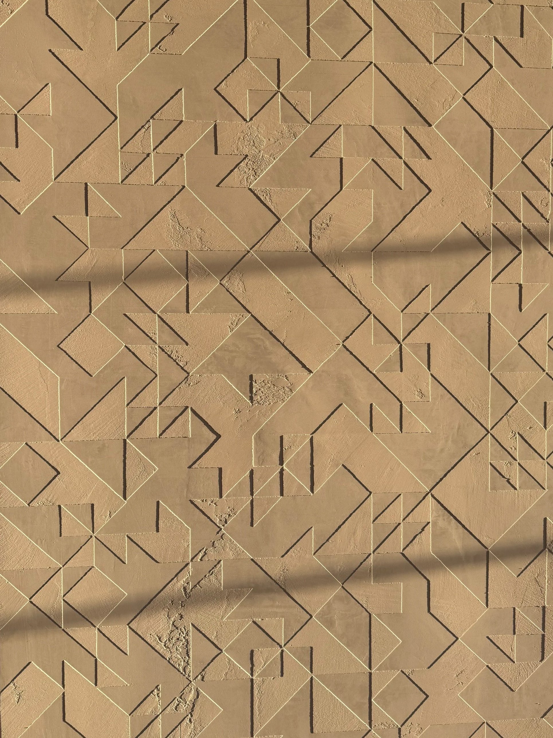 A textured beige wall with geometric patterns and carved shapes, illuminated by sunlight.