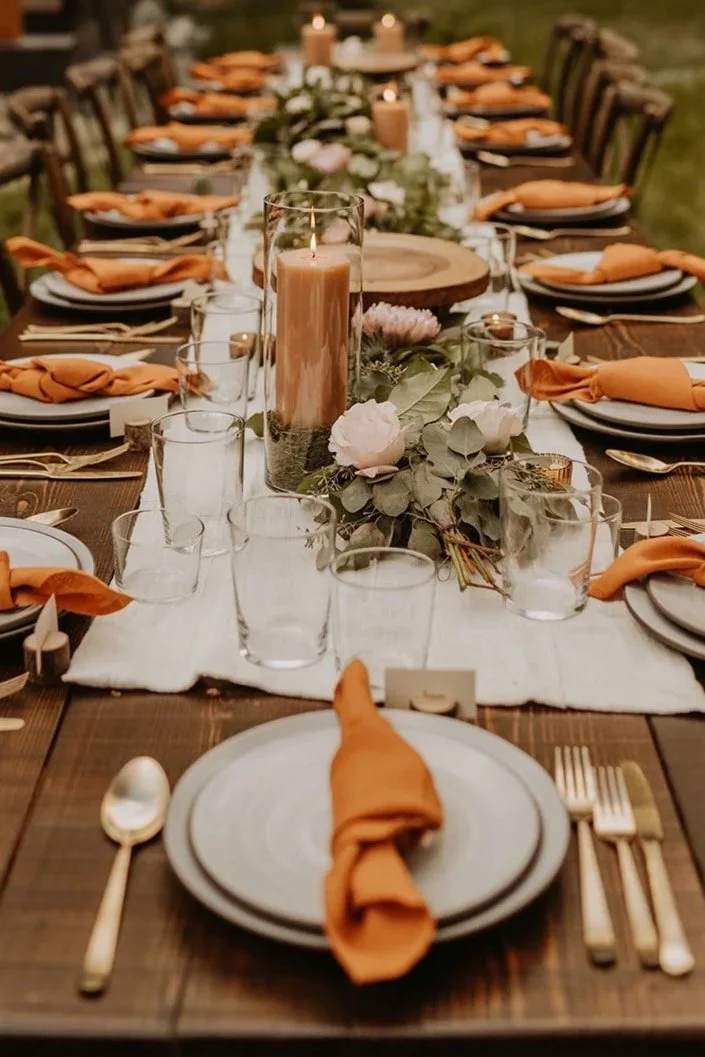 Long outdoor dining table set for a meal, decorated with candles, greenery, and pink flowers, with brown chairs and orange napkins at each place setting, in a wooded area.