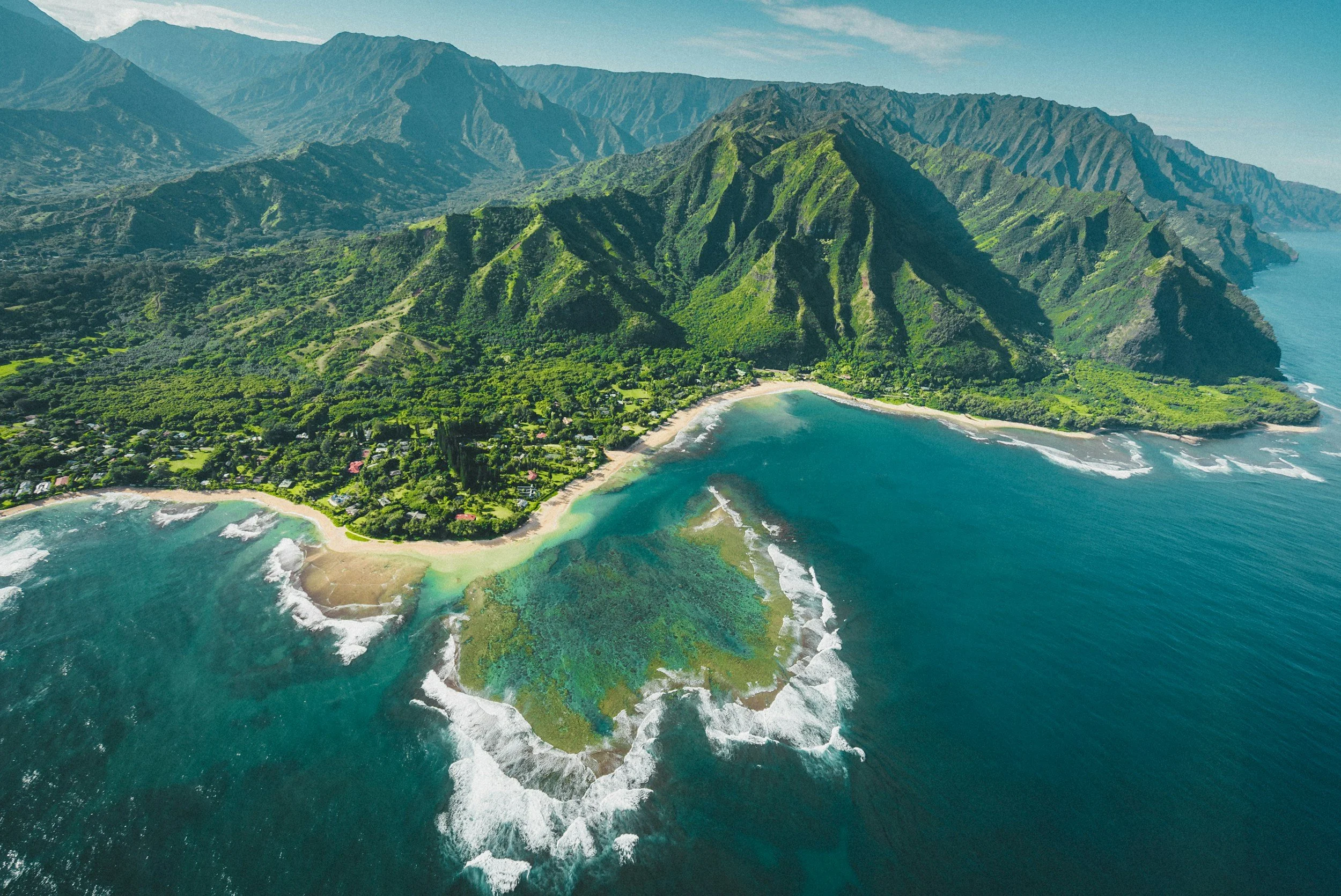 Aerial view of lush green mountains meeting a coastline with sandy beaches and turquoise water, with small waves breaking on the shore.